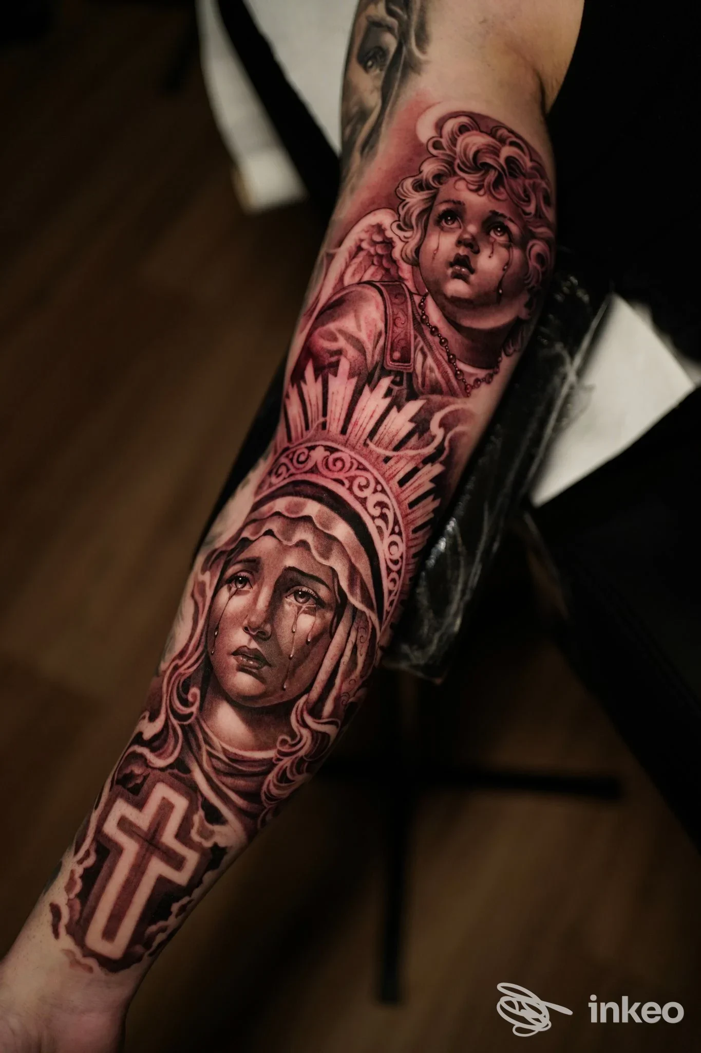 Braccio con tattoo realistico a tema sacro, volti femminili, croce e ornamenti su pelle reale.