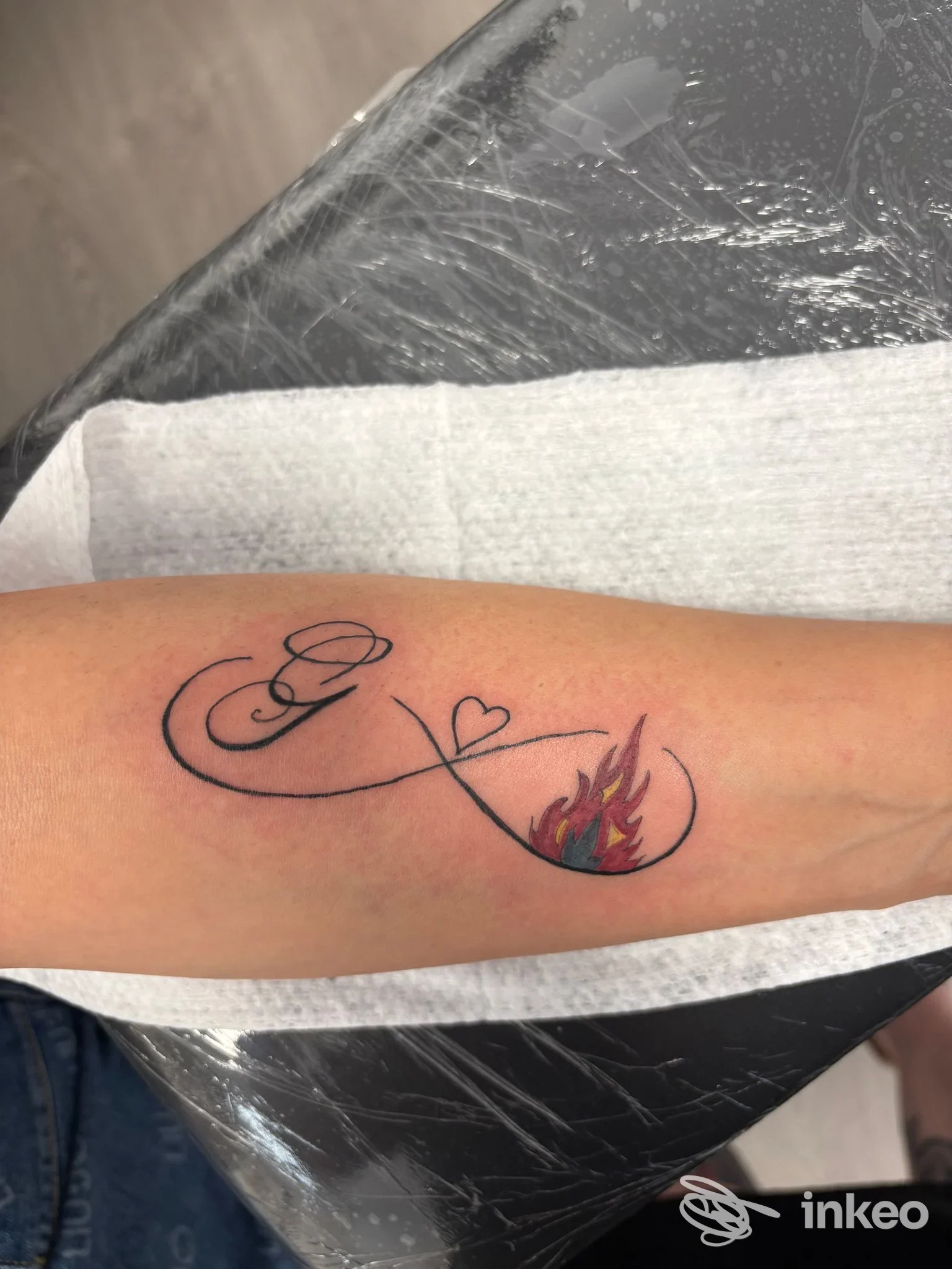Tatuaggio su avambraccio con motivo infinito, cuore e fiamma colorata all’estremità destra, senza presenza di viso. realizado nello studio All Black Tattoo.