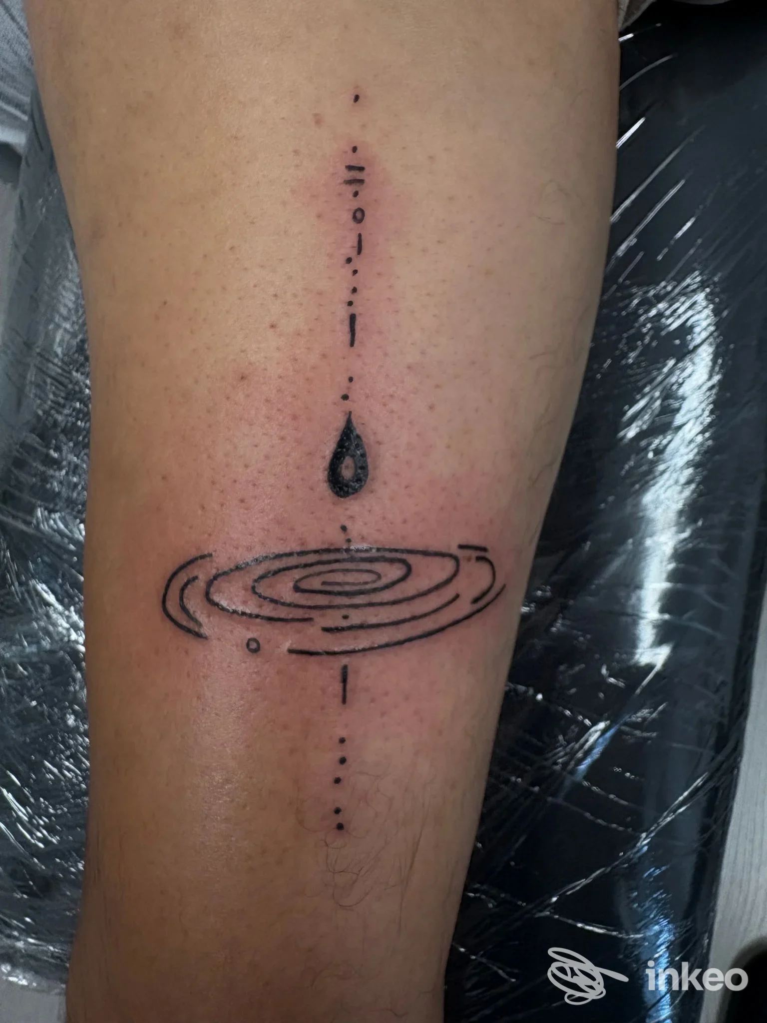 Tatuaggio reale sul braccio raffigurante una linea verticale con puntini, una goccia nera al centro e cerchi concentrici che ricordano onde, realizzato sulla pelle.