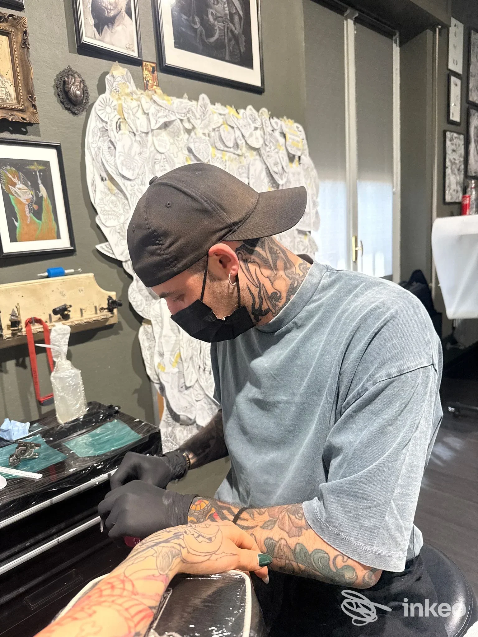 Un tatuatore con mascherina e berretto esegue un tatuaggio sull'avambraccio di un cliente al banco di uno studio a Montichiari, pareti decorate e strumenti visibili.