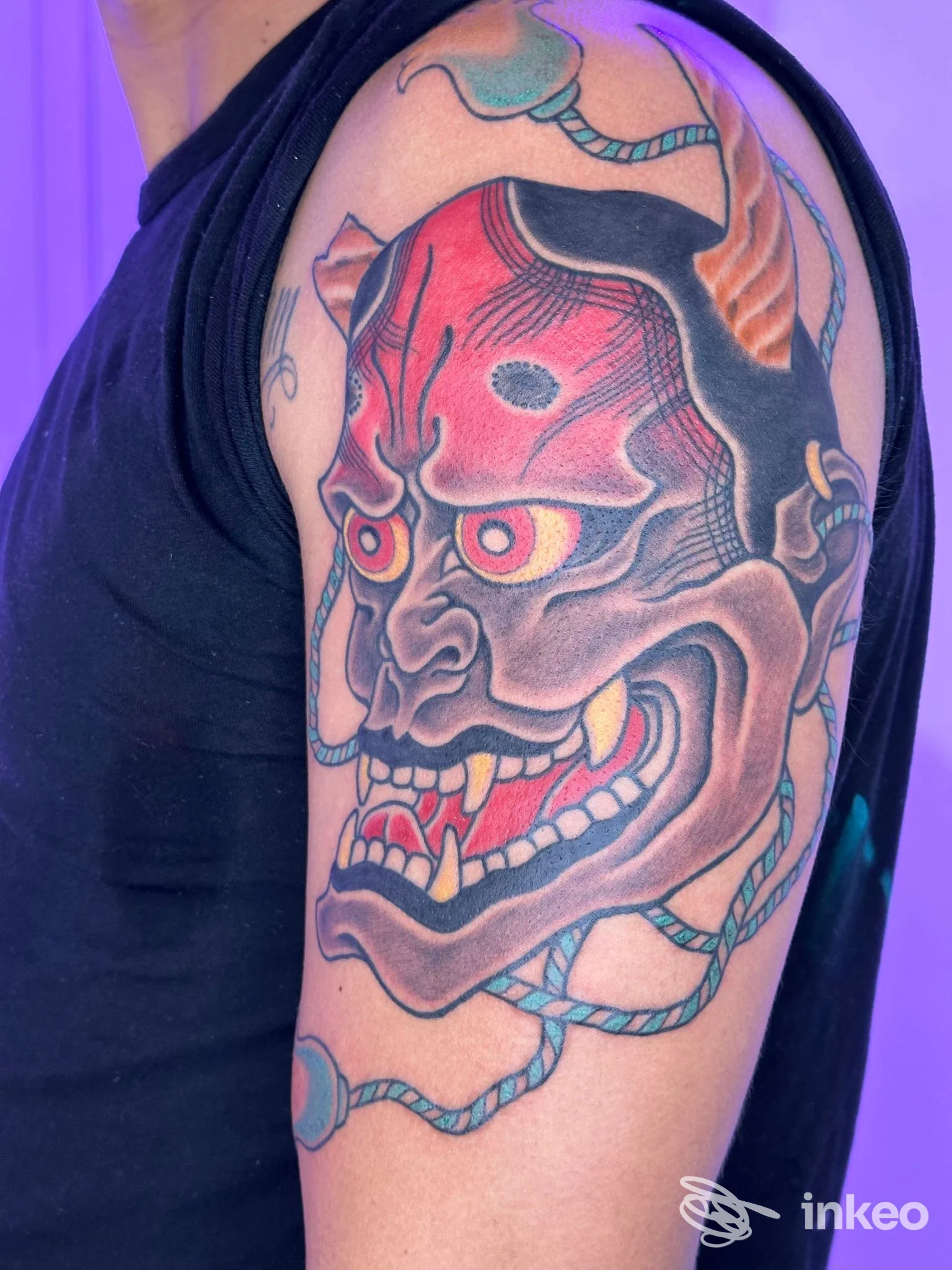 Grande tatuaggio su braccio superiore a Roma realizzato da Baltica Tattoo: maschera demoniaca colorata in rosso, nero e toni teal.