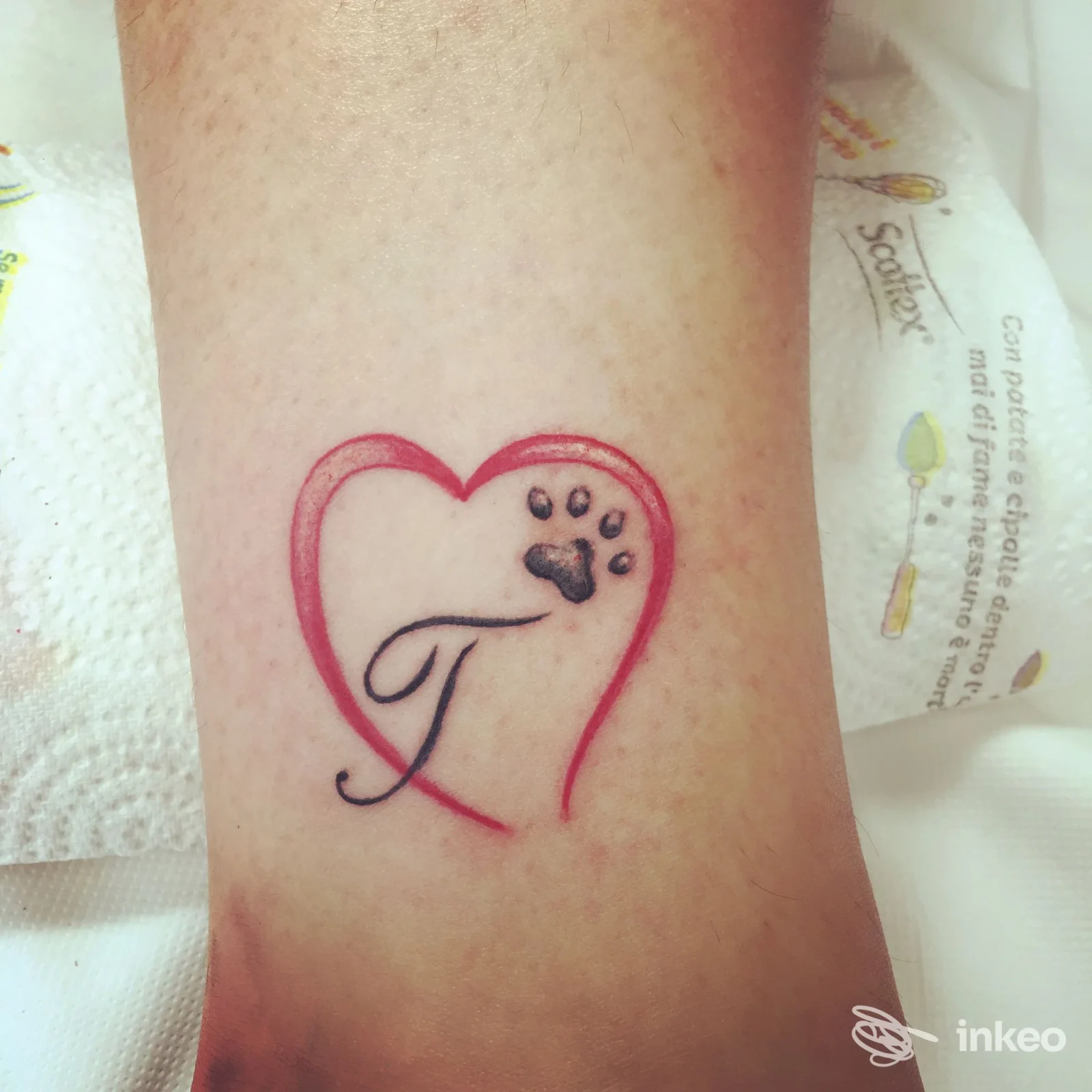 Tattoo su pelle reale di un piccolo cuore rosso con una zampa nera all'interno e una lettera stilizzata, visibile sull’avambraccio.