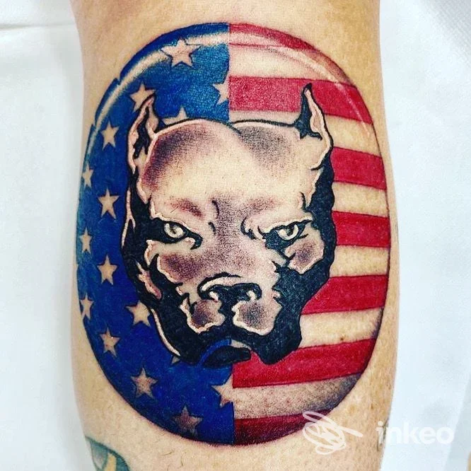Tattoo su pelle raffigurante un cane con sfondo a bandiera USA, ovalo colorato; realizza contorno e colori vividi su braccio.
