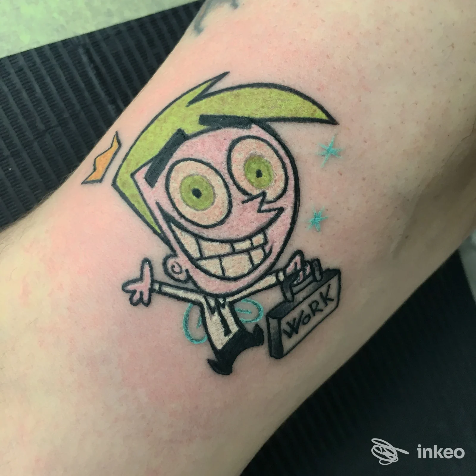 Tattoo colorato su pelle raffigurante un personaggio cartoon con grandi occhi verdi e sorriso, situato sull’avambraccio.