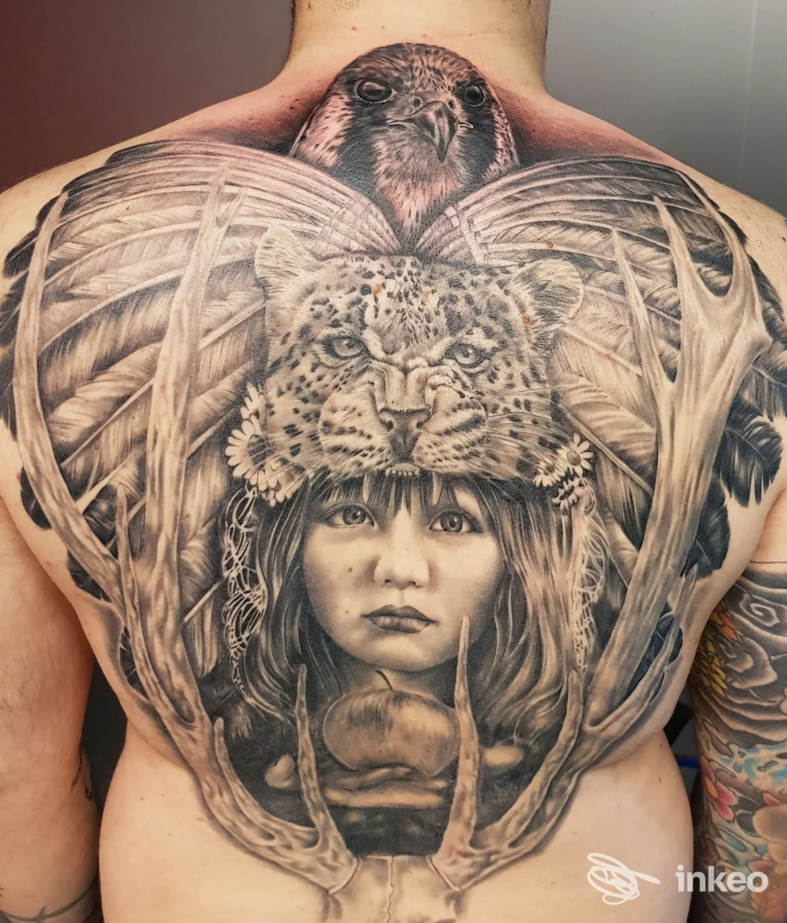 Tattoo realistico su schiena raffigurante figure animale con dettagli in bianco e nero, con cornice di ali e testa centrale; non si vede volto umano completo.