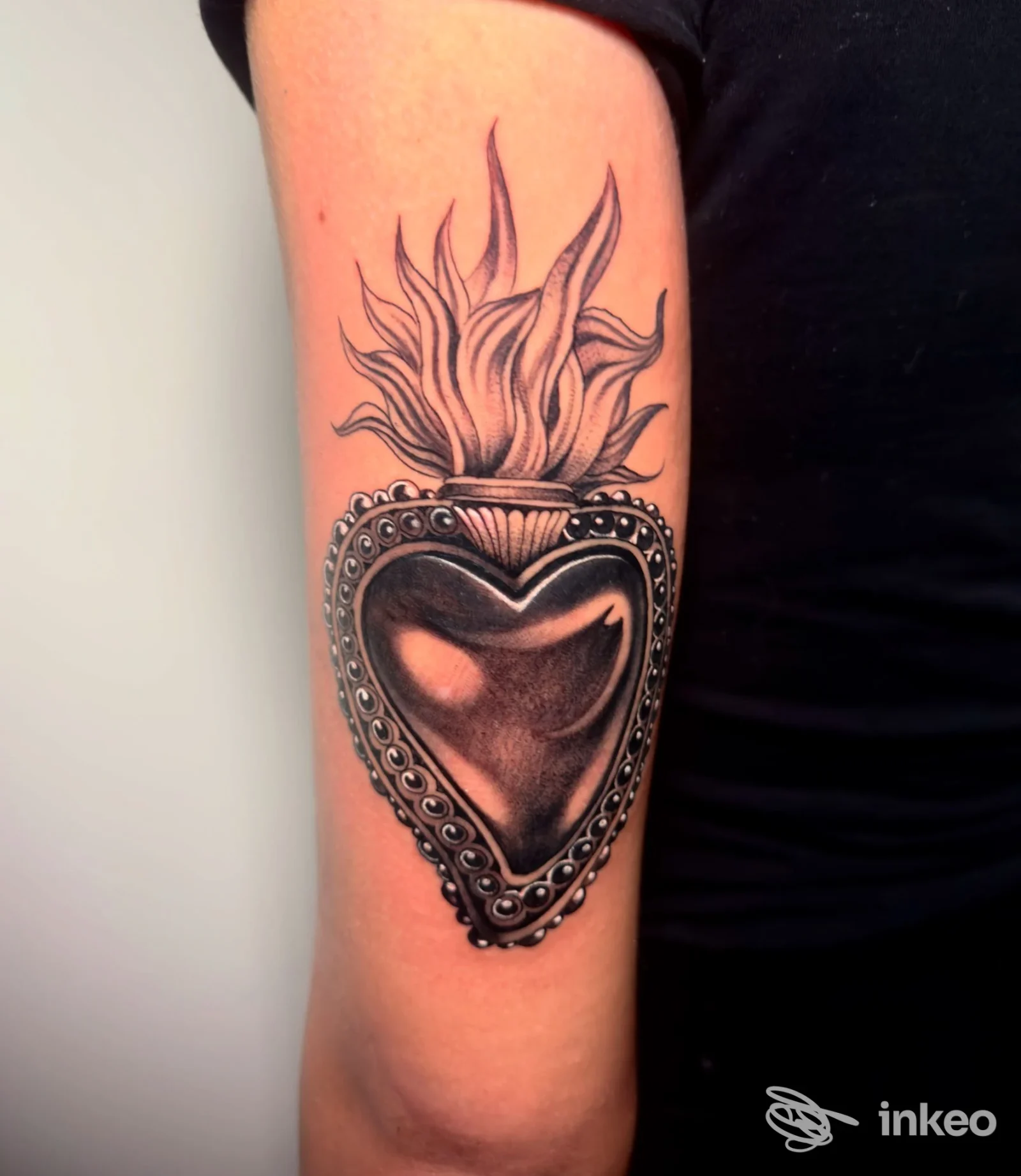Tatuaggio a cuore in bianco e nero con cornice ornamentale e fiamme sul braccio superiore, foto per STORIE DI VITA SU PELLE, Fossano.