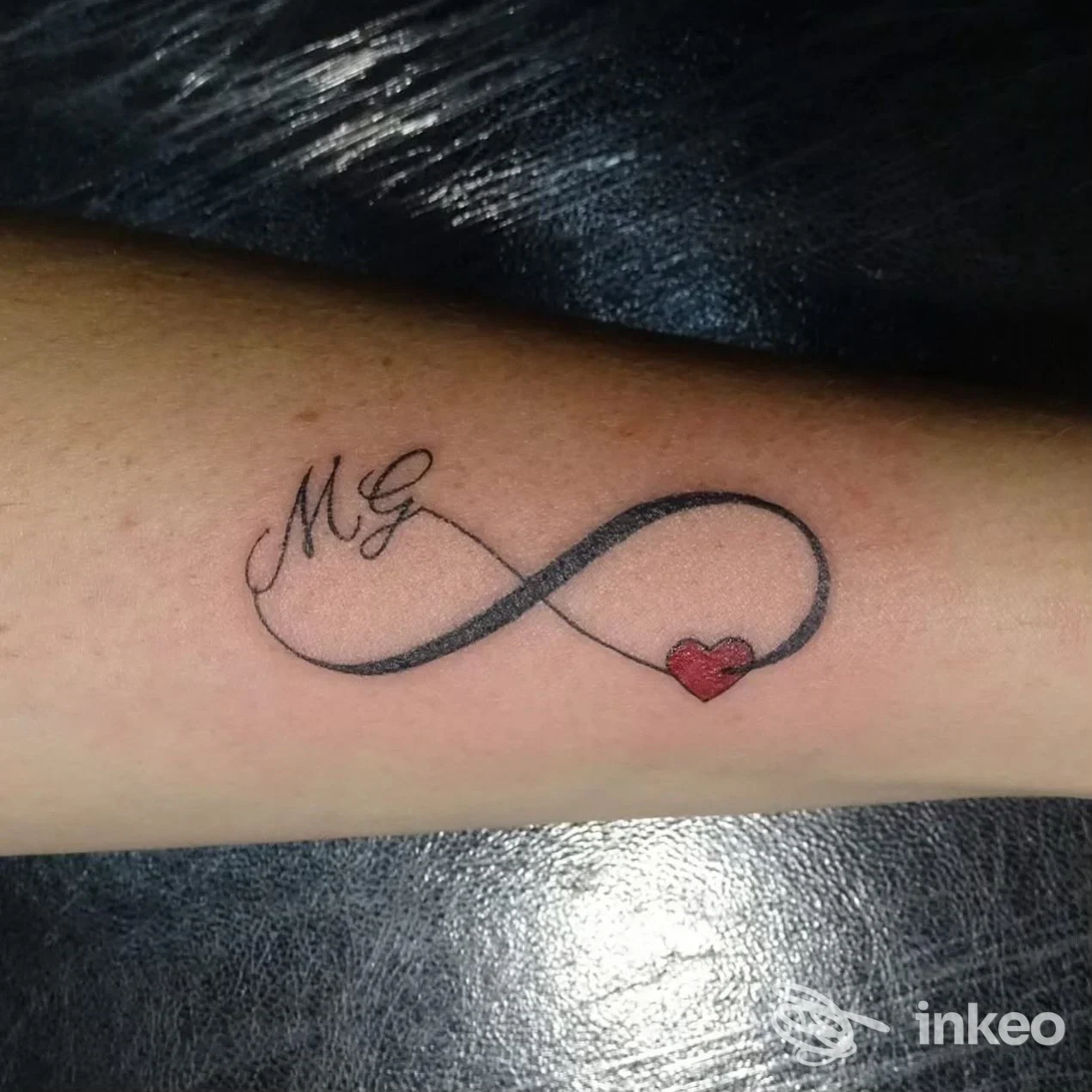 Tatuaggio infinito nero con cuore rosso e iniziali 'M G' sull'avambraccio, realizzato da Barraco Design a Marsala.