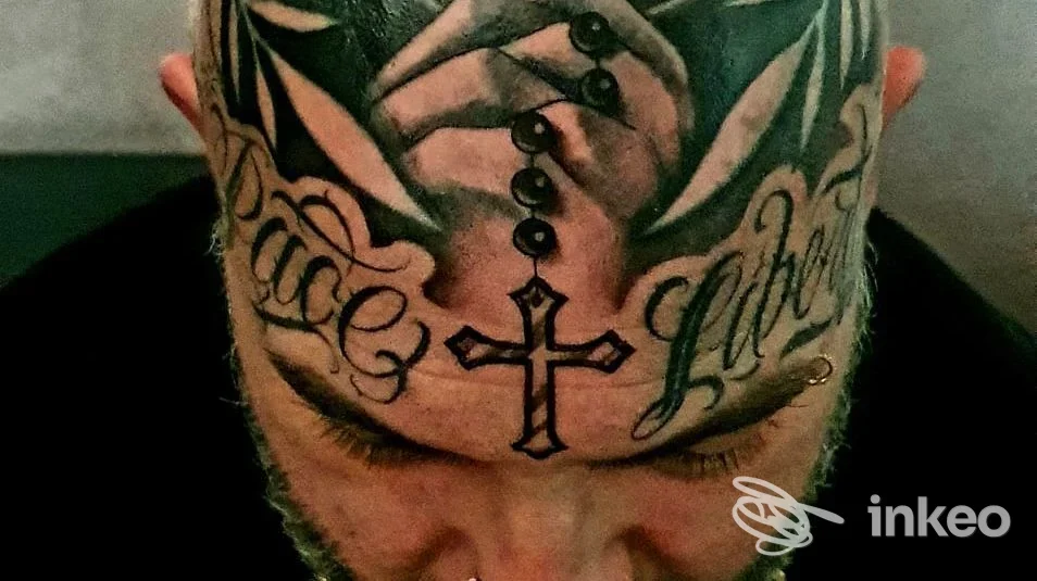 Primo piano della fronte tatuata con croce centrale, scritte ornamentali laterali e motivi scuri, lavoro su pelle per Hd piercing -tatuaggi Family Olbia, Olbia.