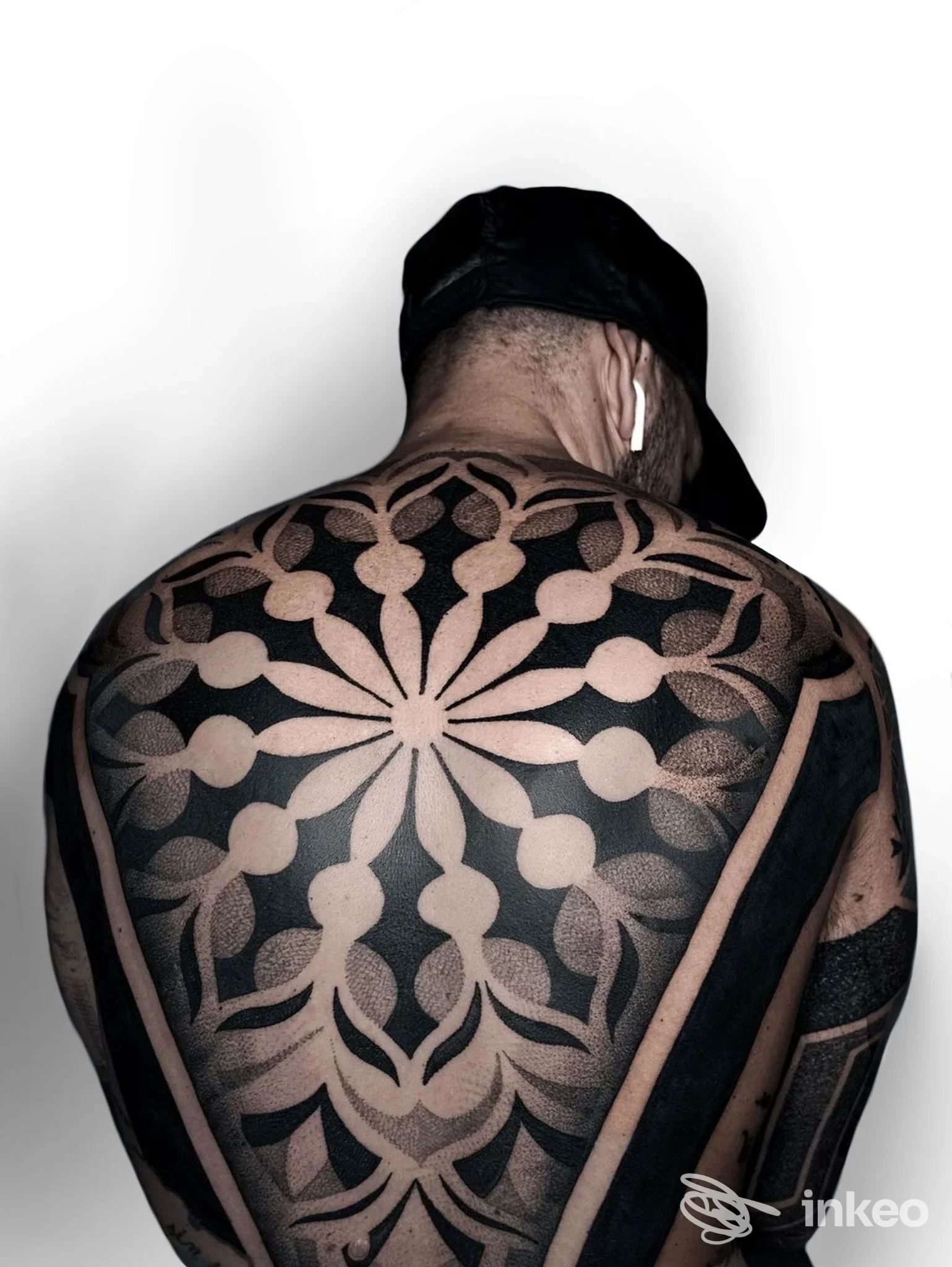 Tattoo molto esteso sulla schiena di una persona, disegno circolare geometrico in tonalità scure, visibile dal retro con cappuccio. nessun volto intero visibile.