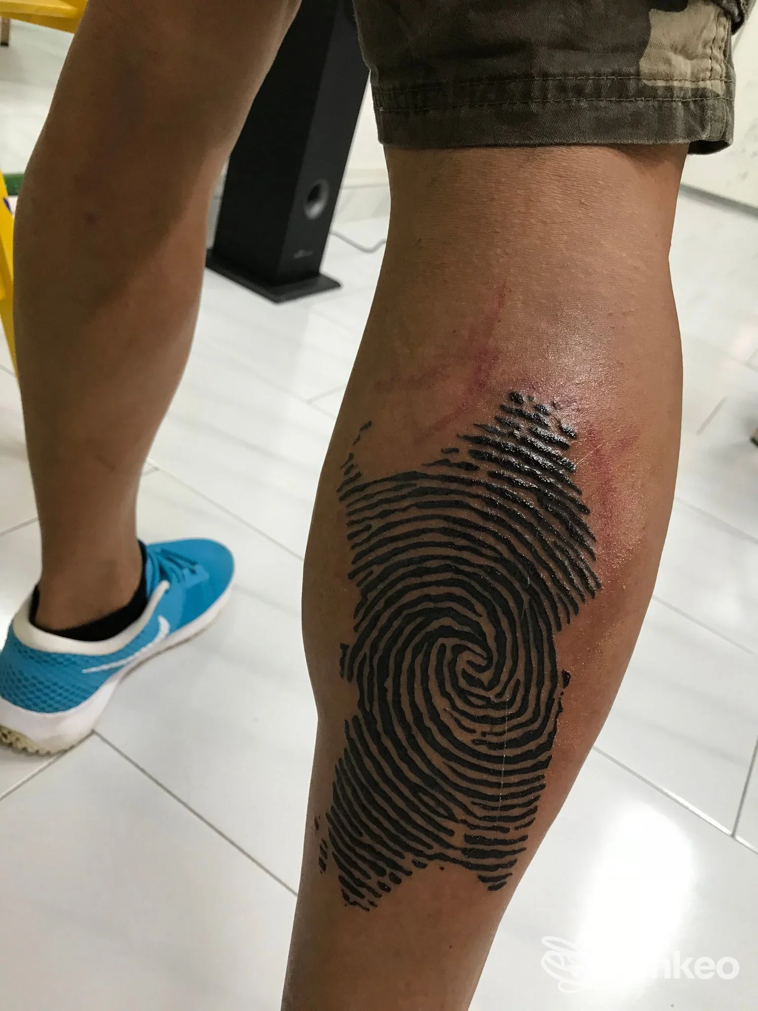 Tatuaggio a impronta digitale sul polpaccio destro fotografato in studio da Vespink Tattoo Studio a Carbonia, primo piano su gamba e pavimento piastrellato.