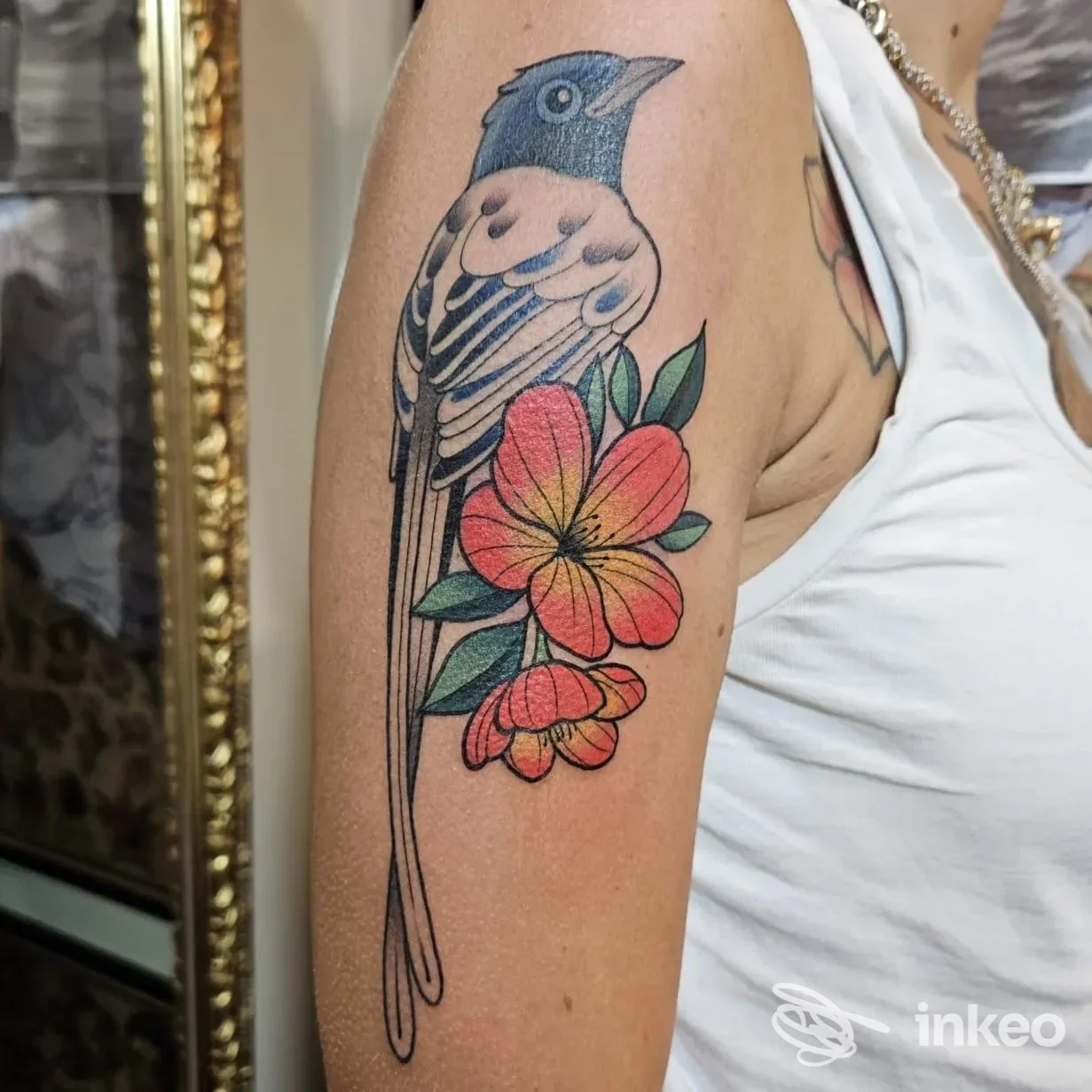 Tattoo a tema uccello azzurro e fiori rosa-arancione sull’avambraccio, colori vivaci e linee pulite, visibile parte del braccio con sfondo morbido.