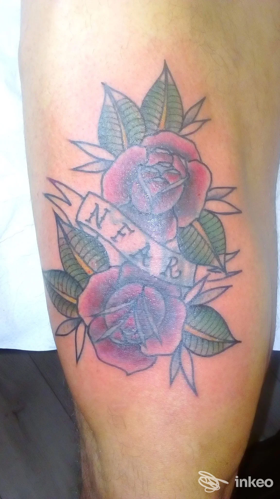 Tattoo a colori su pelle di gamba: due rose rosse con foglie verdi, attraversate da un nastro con testo non pienamente leggibile.