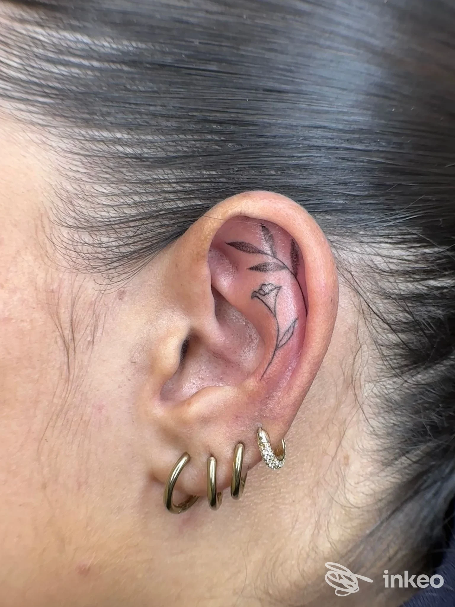 Dettaglio di un tatuaggio minimal a linee nere nella conca dell'orecchio, eseguito da Think Giulia Ghini a Poggibonsi; foto in primo piano mostra anche piccoli orecchini.