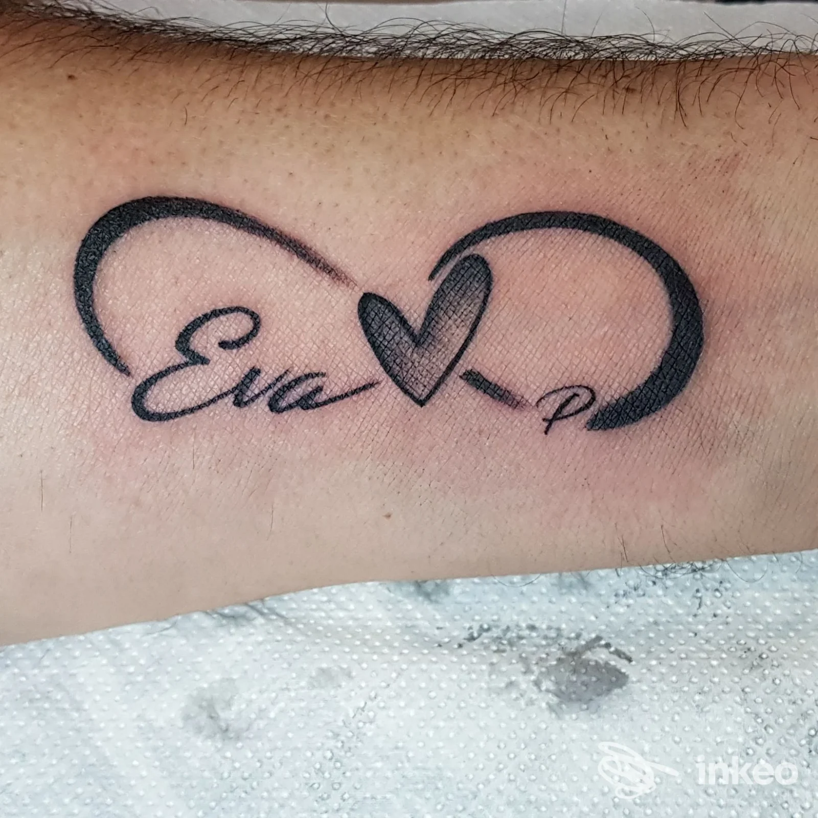 Tattoo reale sull'avambraccio raffigurante un infinito con cuore al centro e testo in corsivo ai lati; colore nero su pelle chiara.