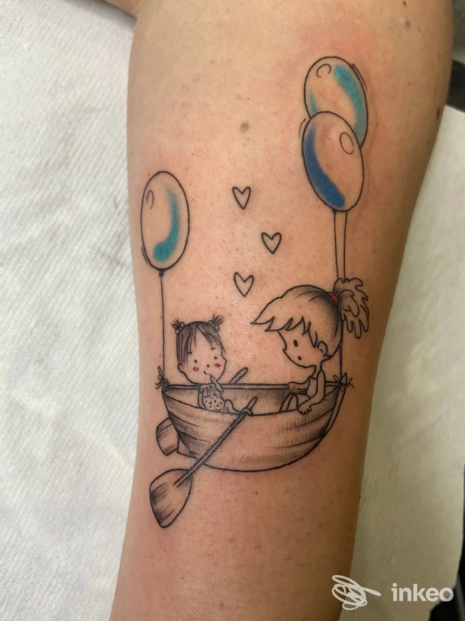 Tattoo su braccio raffigurante due bambini in una barca, con palloncini azzurri, stile cartone animato, tratto nero ben definito e colori riempitivi.
