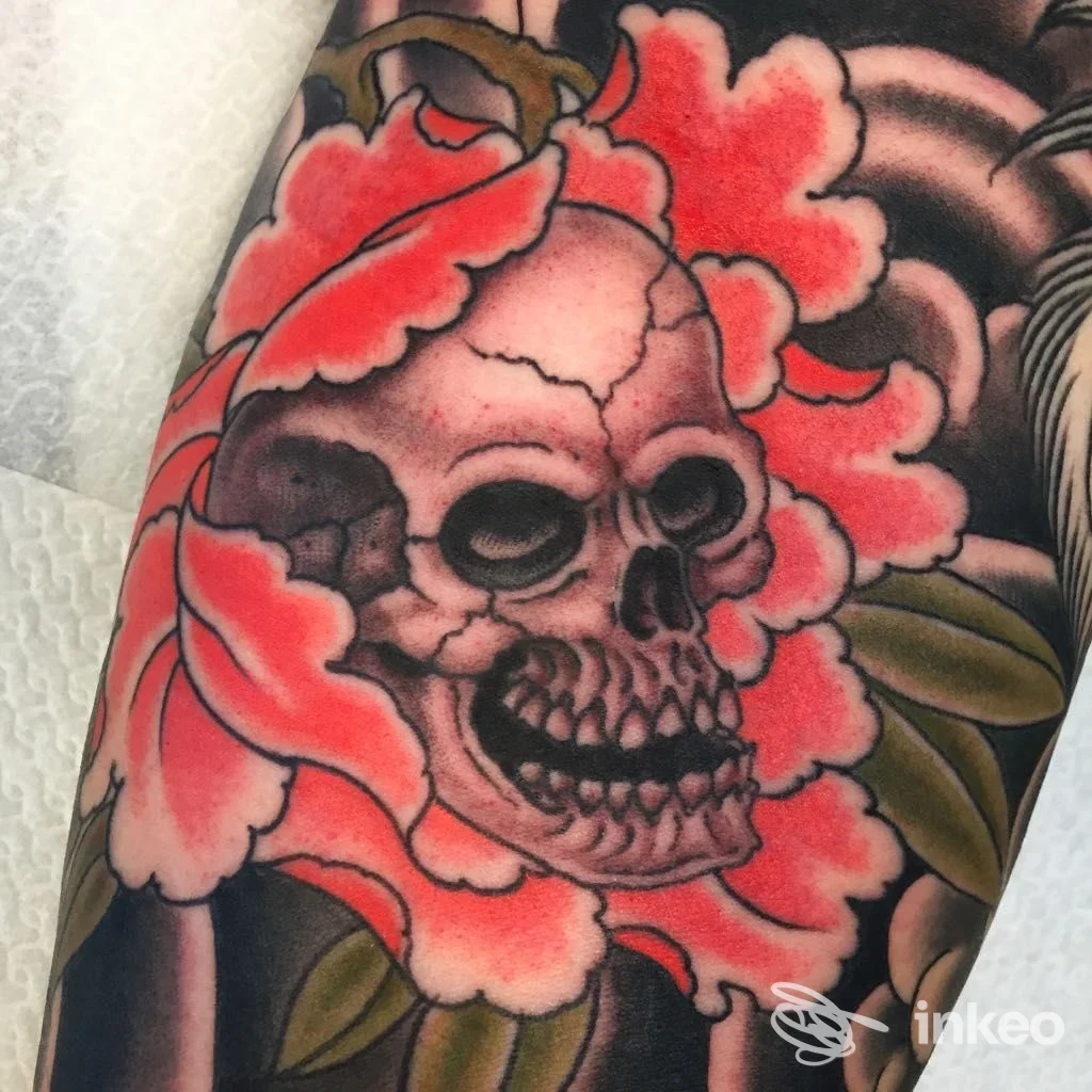 Dettaglio di tatuaggio su braccio: teschio centrale circondato da petali rossi e foglie verdi, lavoro fotografato da Tattoo River a Vicenza.