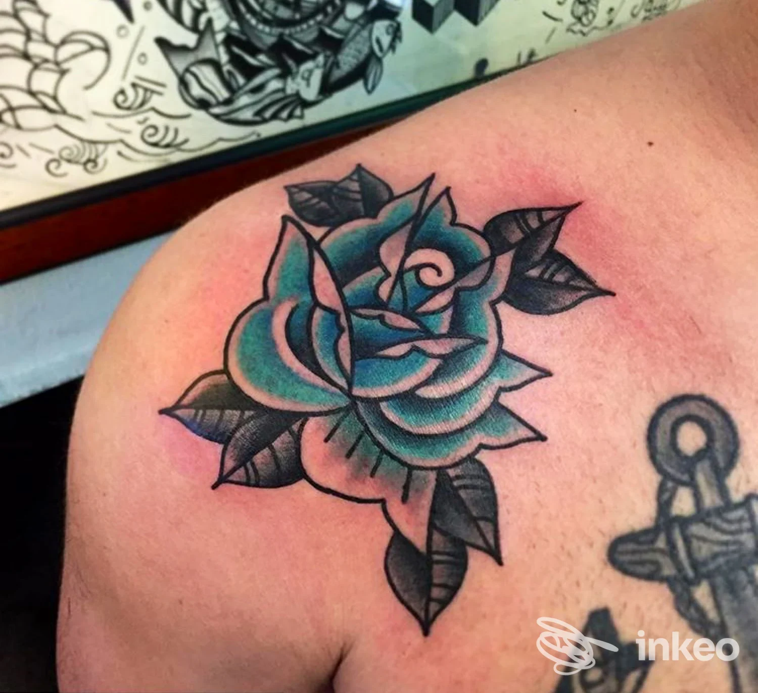 Tatuaggio a rosa azzurra su spalla, linea nera marcata e sfumature blu-verdi, con foglie scure; pedina un piccolo tratto di tatoo vicino. Nessun testo o interfaccia visiva presente.