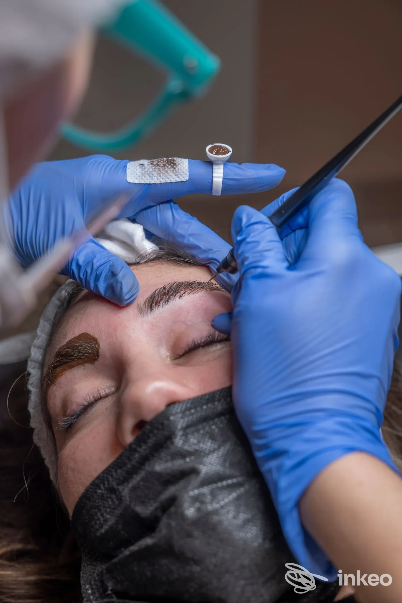 Operatrice di Valentina Beauty Studio a Verona esegue dermopigmentazione sopracciglia su cliente con occhi chiusi, guanti blu e mascherina nera.