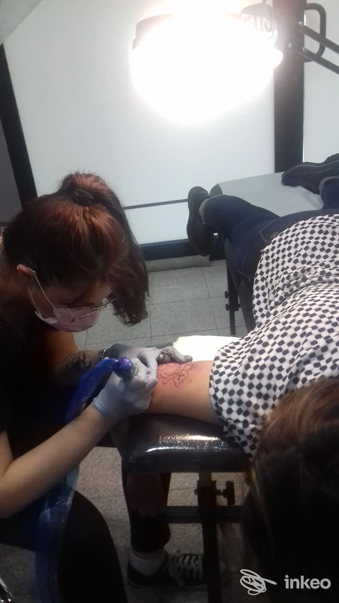 In uno studio di Cassano Magnago, Black Cat Tattoo, un’operatrice sta realizzando un tatuaggio sul braccio di un cliente, con staff medico e luce da studio.