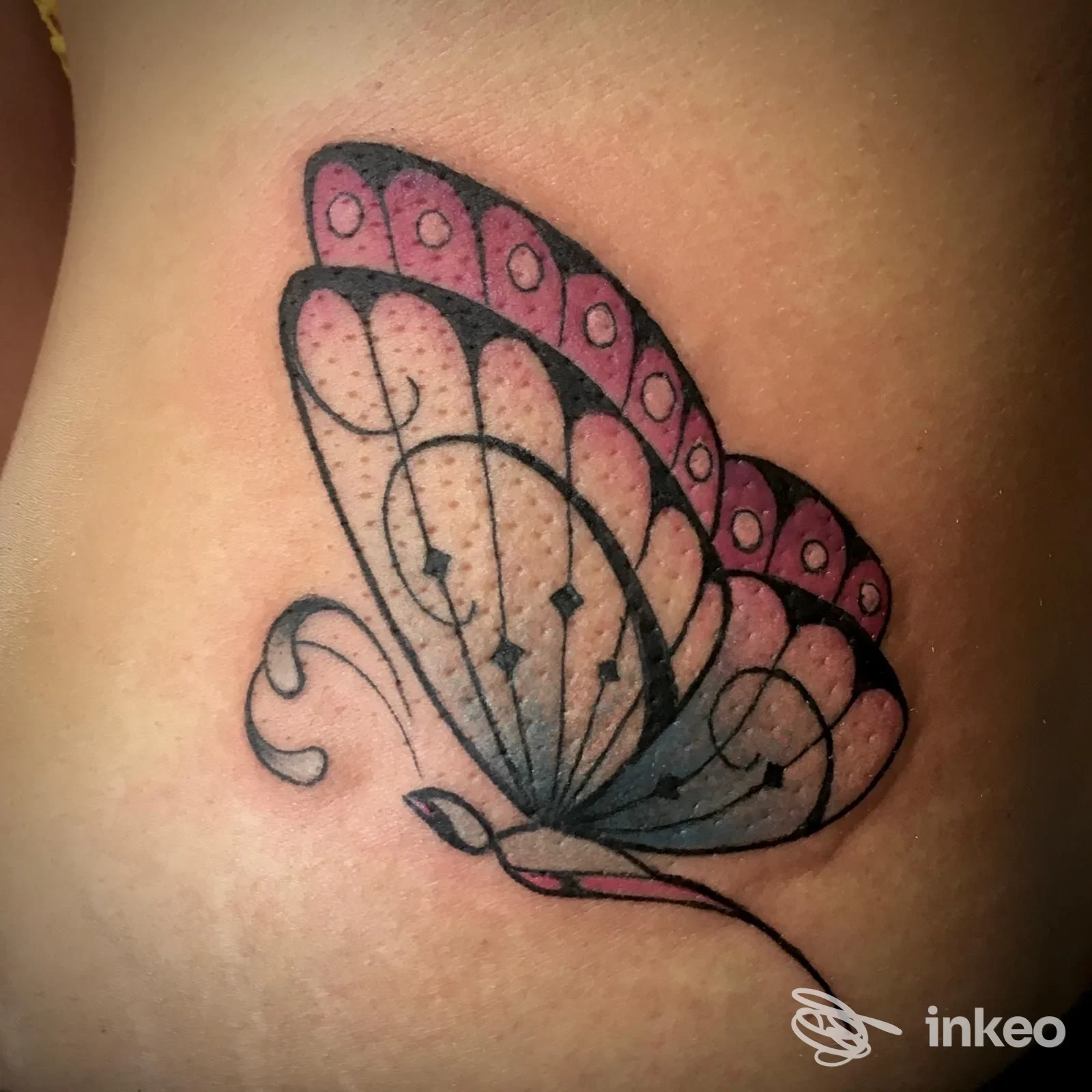 Tattoo di una farfalla rosa e blu sul fianco, eseguito da SONOS TATTOO ART ad Arbatax, fotografia ravvicinata della pelle.