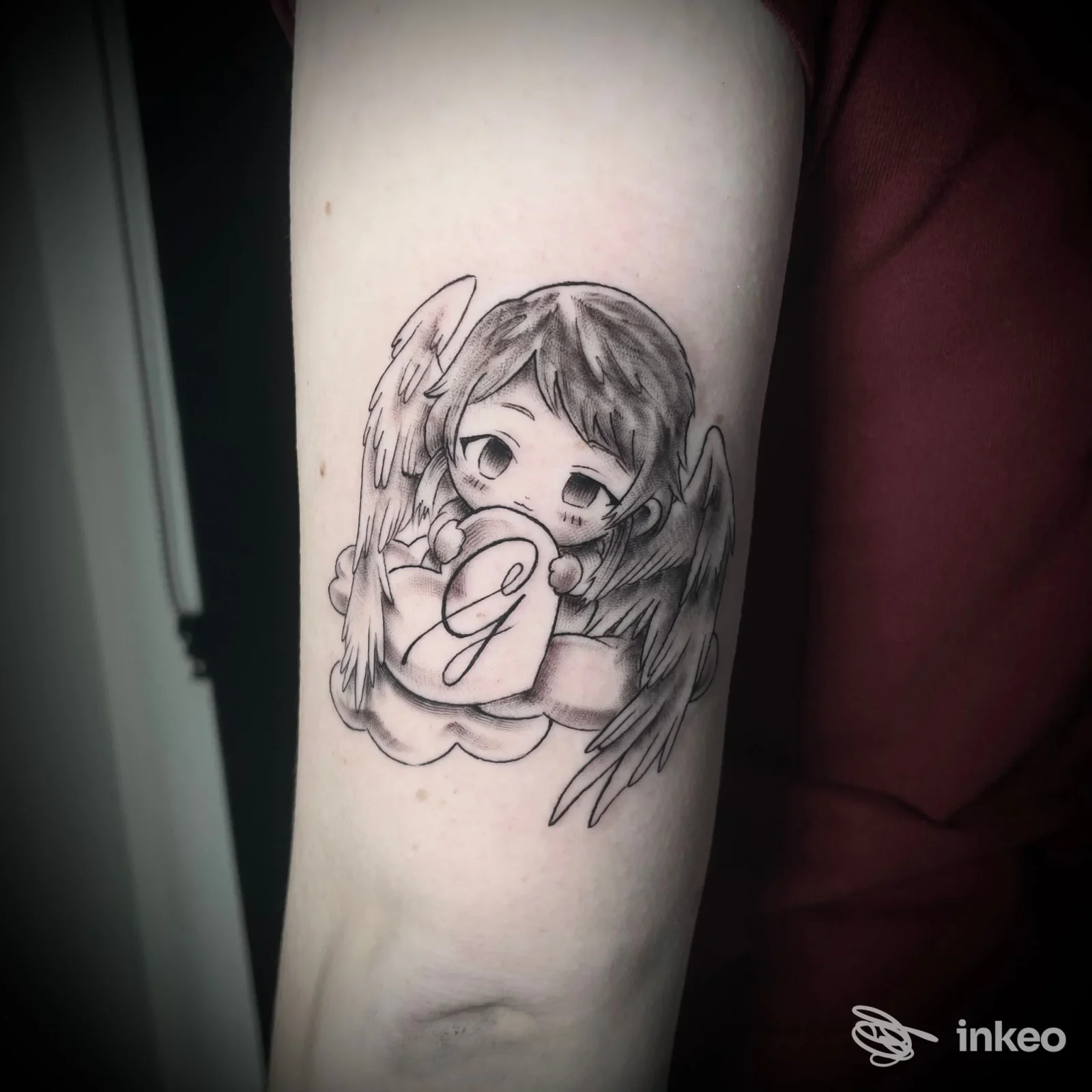 Tatuaggio in stile chibi a inchiostro nero su avambraccio: angelo che tiene un cuore con la lettera G. Al di là dello Specchio Tattoo Studio, Saccon.