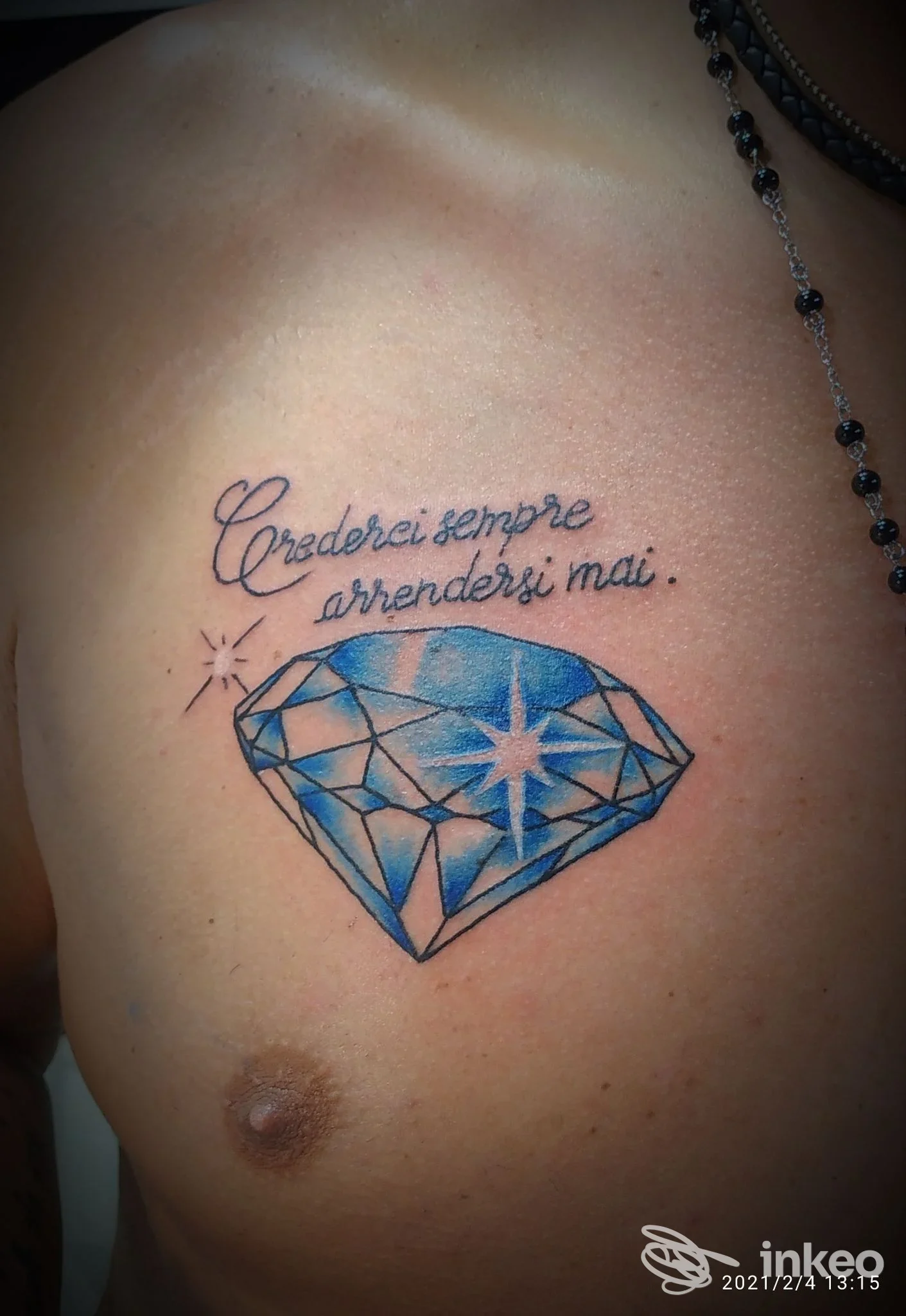 Tatuaggio sul petto con diamante blu e scritta corsiva 'Crederci sempre arrendersi mai.' Realizzato da Micky Tattoo Studio a Casalecchio di Reno.