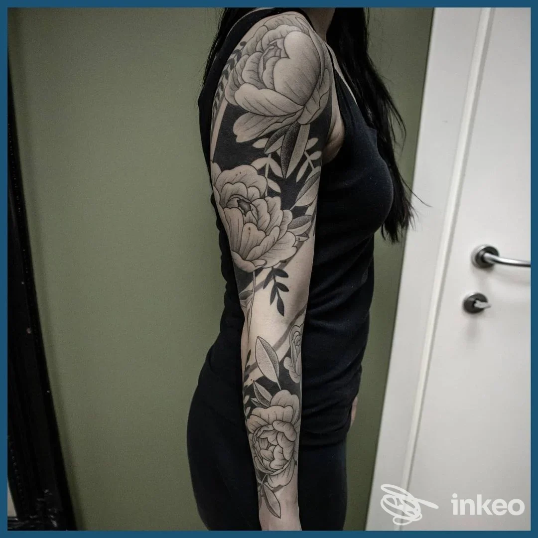 Donna di profilo mostra un braccio sinistro completamente tatuato con motivi floreali in bianco e nero; indossa una maglietta nera, sfondo interno con porta visibile.
