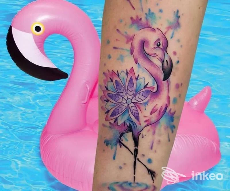 Tatuaggio colorato su avambraccio raffigurante un fenicottero rosa con fiore e schizzi blu, sullo sfondo una piscina, realizzato su pelle umana.