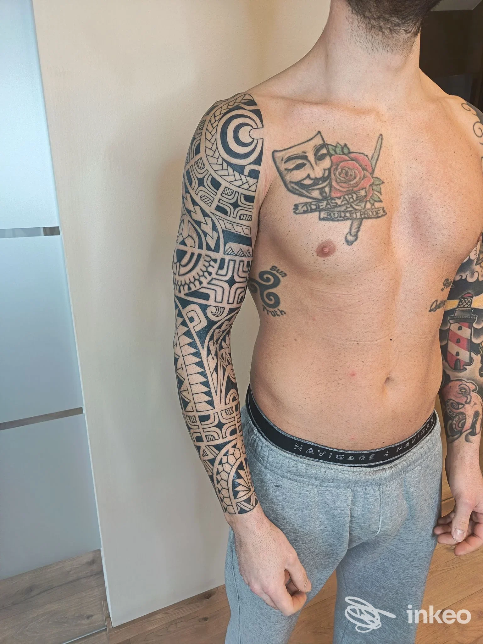 Uomo senza camicia mostra braccio sinistro completamente tatuato e torace con tattoo; nessun overlay visibile.