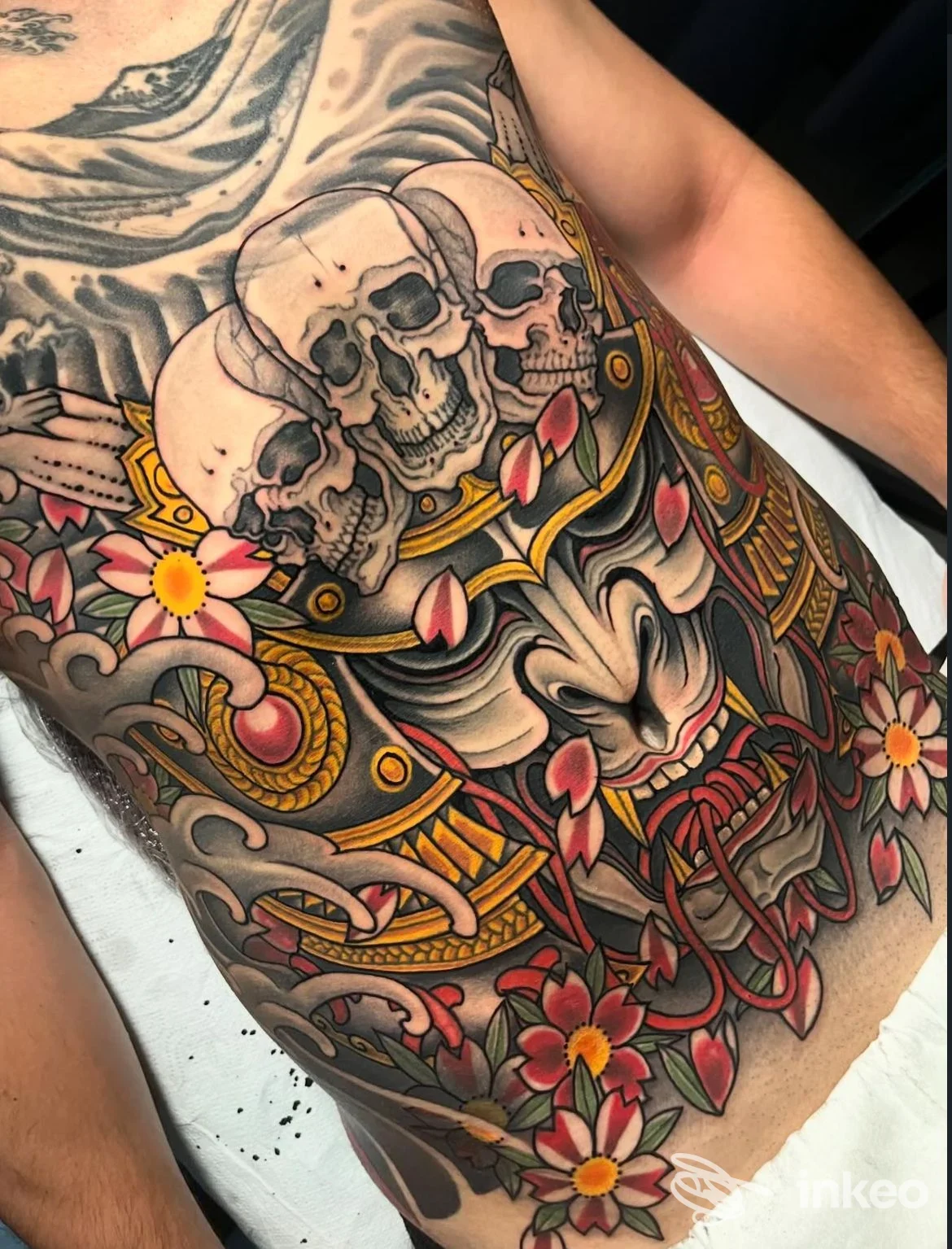Tattoo ampia su torace e fianco sinistro con teschi sovrapposti, ornamenti dorati e fiori rosa. colori vividi e linee nette, visibile parte di braccio e pelle nuda.