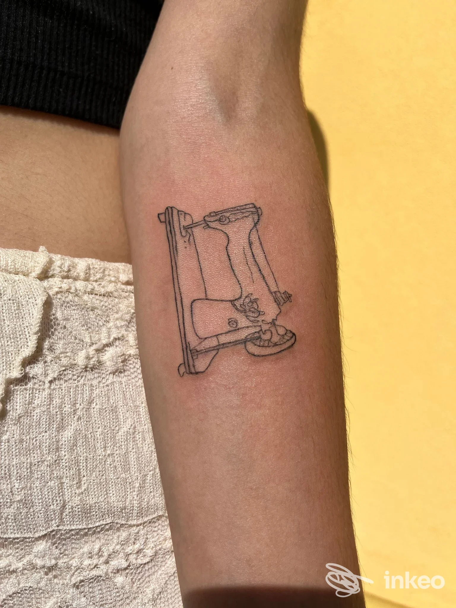 Tattoo su pelle di un braccio raffigurante una macchina da cucire, disegno lineare sobrio, senza volto visibile, sfondo giallo.