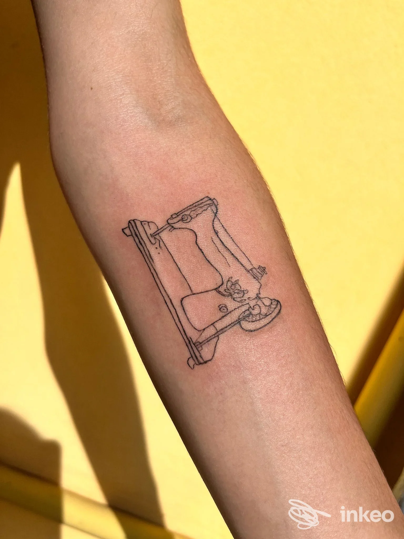 Tatuaggio in line art su avambraccio raffigurante una macchina da cucire, eseguito su pelle; sfondo giallo.