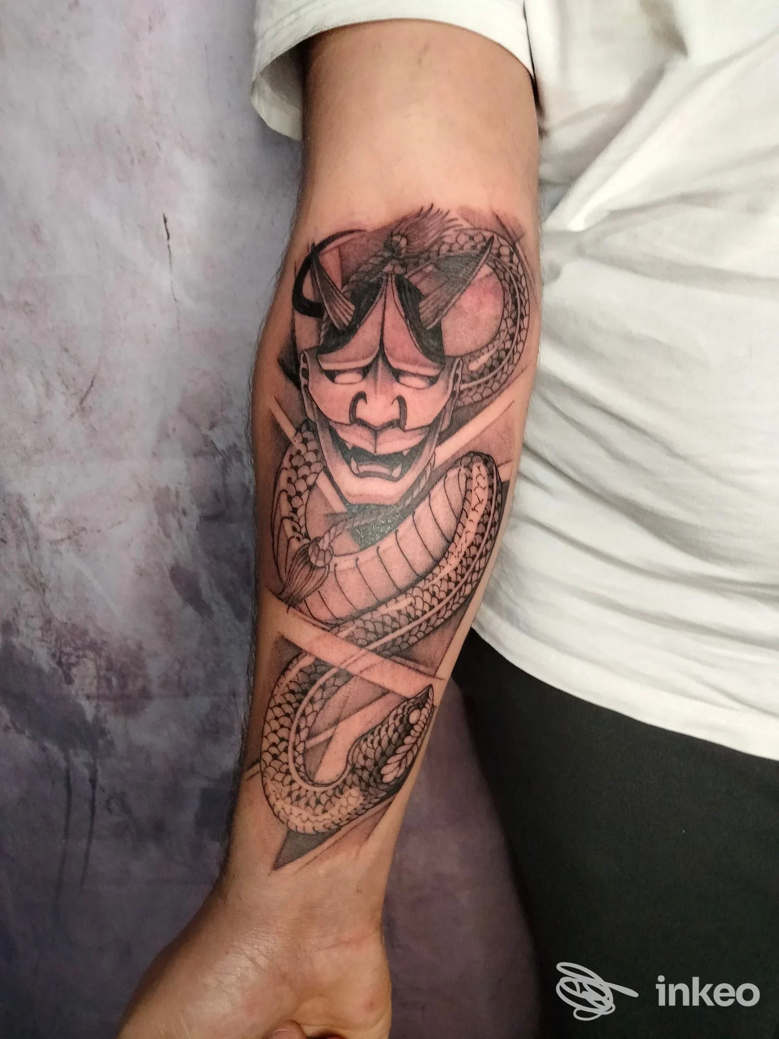 Avambraccio visibile con tatuaggio in bianco e nero raffigurante una maschera stilizzata e un serpente che si avvolge lungo l’avambraccio.