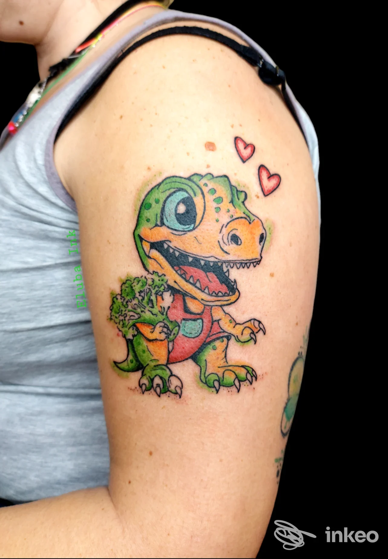Tatuaggio sul braccio sinistro raffigurante un dinosauro cartoonesco colorato con due cuori accanto; l’immagine mostra la pelle e la texture della pelle, senza volto umano visibile.