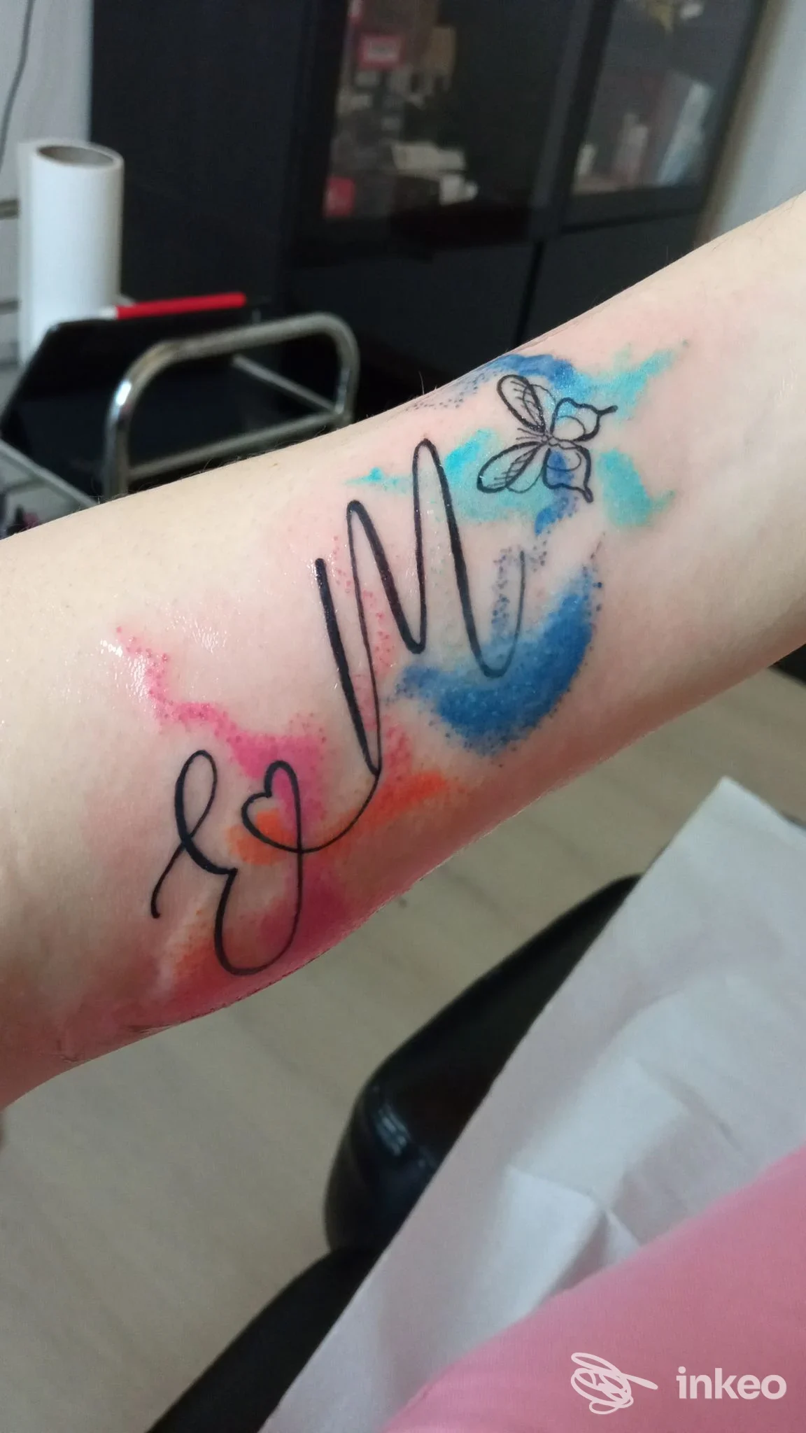 Braccio con tatuaggio acquerello: schizzi colorati blu, rosa e arancione con linee nere che disegnano un cuore e una piccola farfalla.