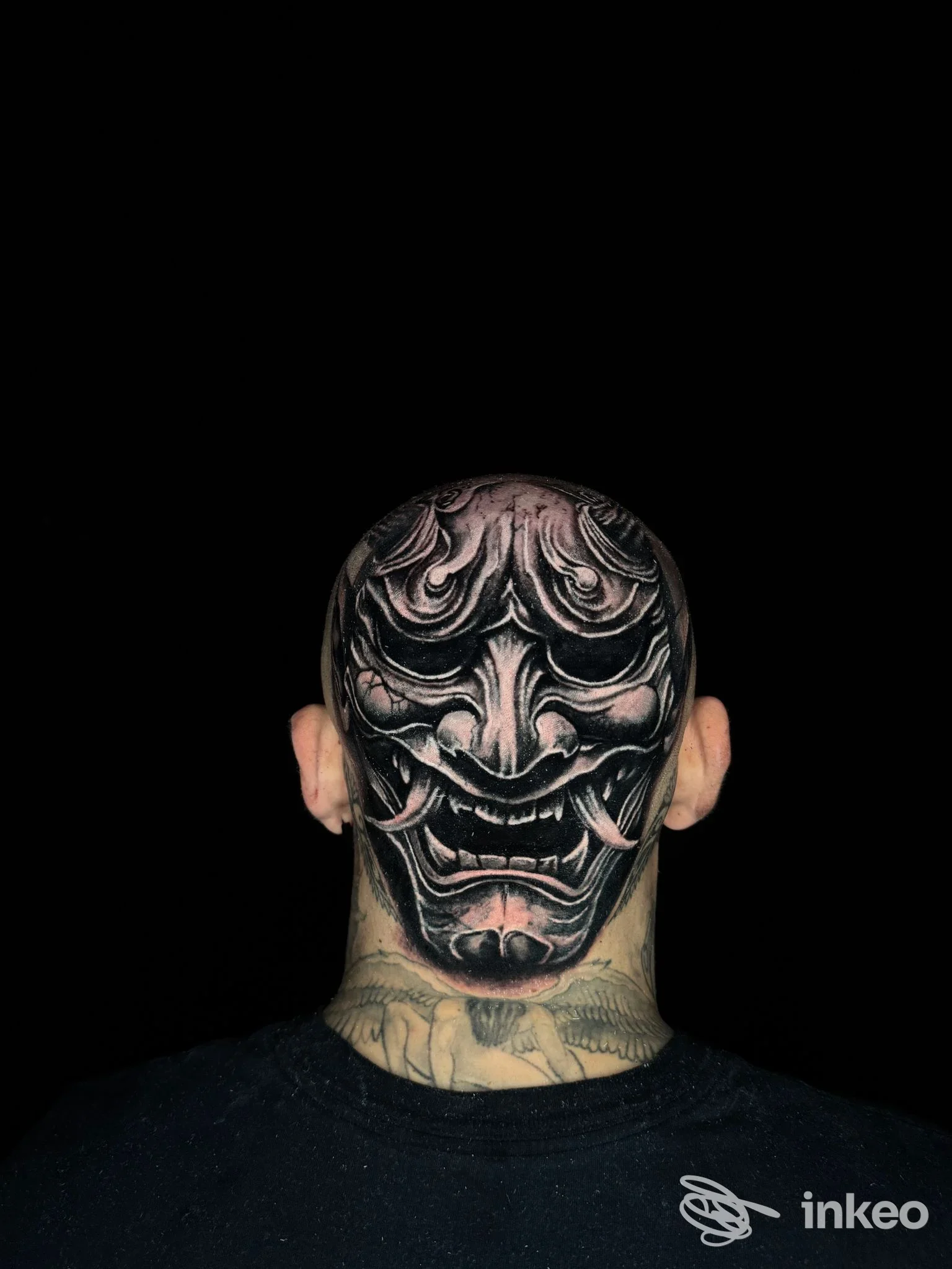 Testa di uomo vista di dietro con tatuaggio nero grigio su cuoio capelluto e collo, raffigurante una maschera demonica, sfondo nero