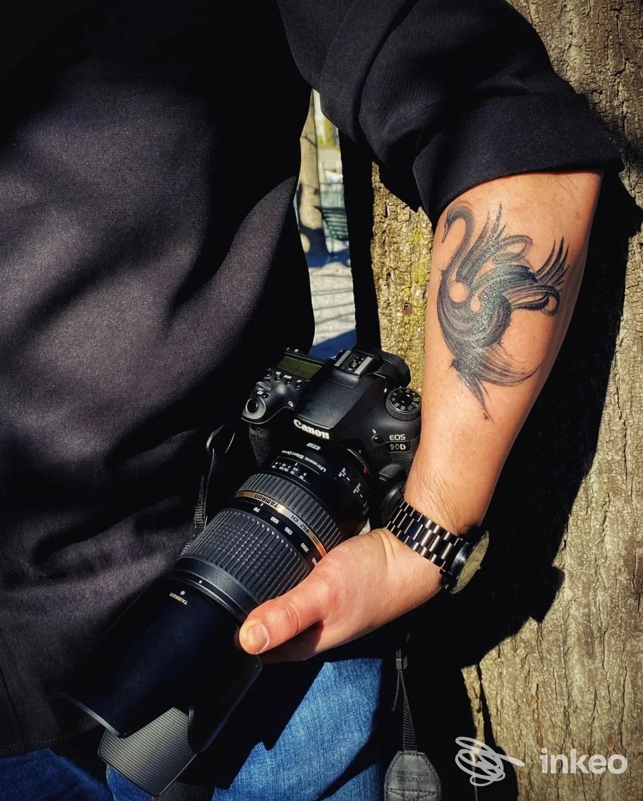 Braccio di persona con tatuaggio stilizzato di uccello o motivo floreale, tenendo una fotocamera reflex con cinturino al polso.