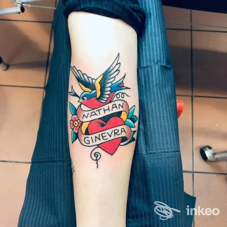 Braccio di una persona con tatuaggio a colori: un cuore al centro con una freccia, una colomba e una fascia con i nomi Nathan e Ginevra; contorni neri e fiori.