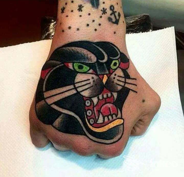 Tattoo a dorso della mano raffigurante una pantera nera con occhi verdi e bocca aperta, colori vivaci, visibile su una mano poggiata su un lenzuolo bianco.
