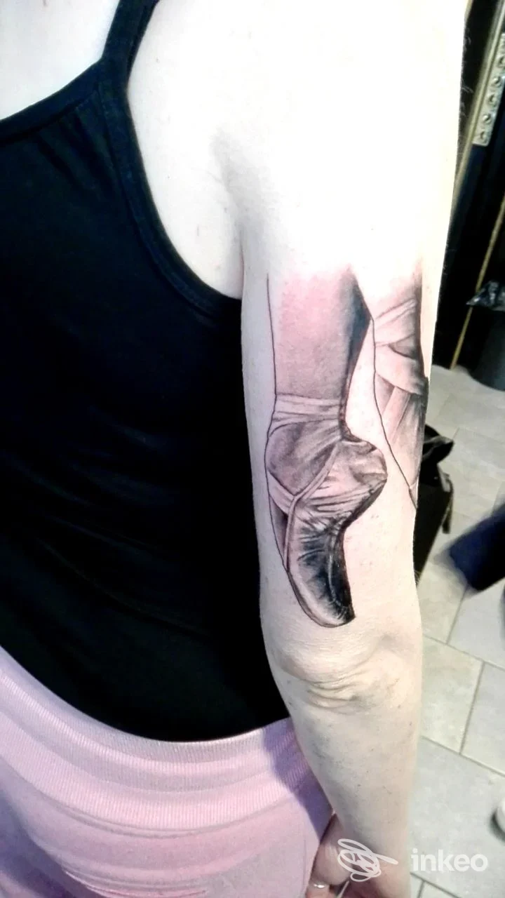 Tattoo visibile sull’avambraccio raffigurante una scarpa alta, realizzato sulla pelle; senza volto visibile, immagine singola, illuminazione neutra, abbigliamento nero sullo sfondo.