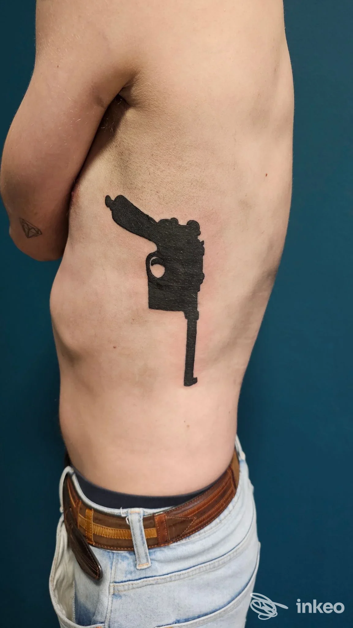 Tattoo nero a forma di pistola stilizzata sulla parte laterale superiore del braccio di una persona, visibile di profilo; jeans e cintura sono visibili.
