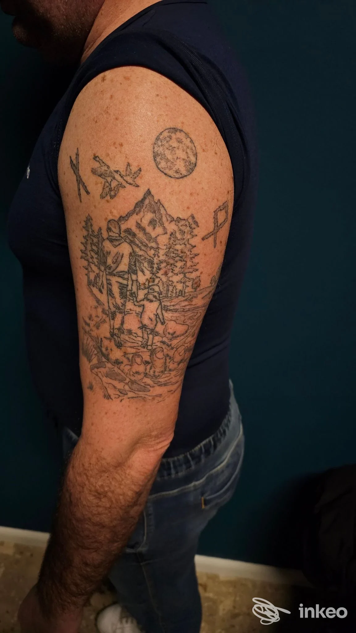 Braccio maschile con tatuaggio visibile: scena all'aperto con montagne, alberi e figura umana, luna piena nel cielo; sfondo scuro.