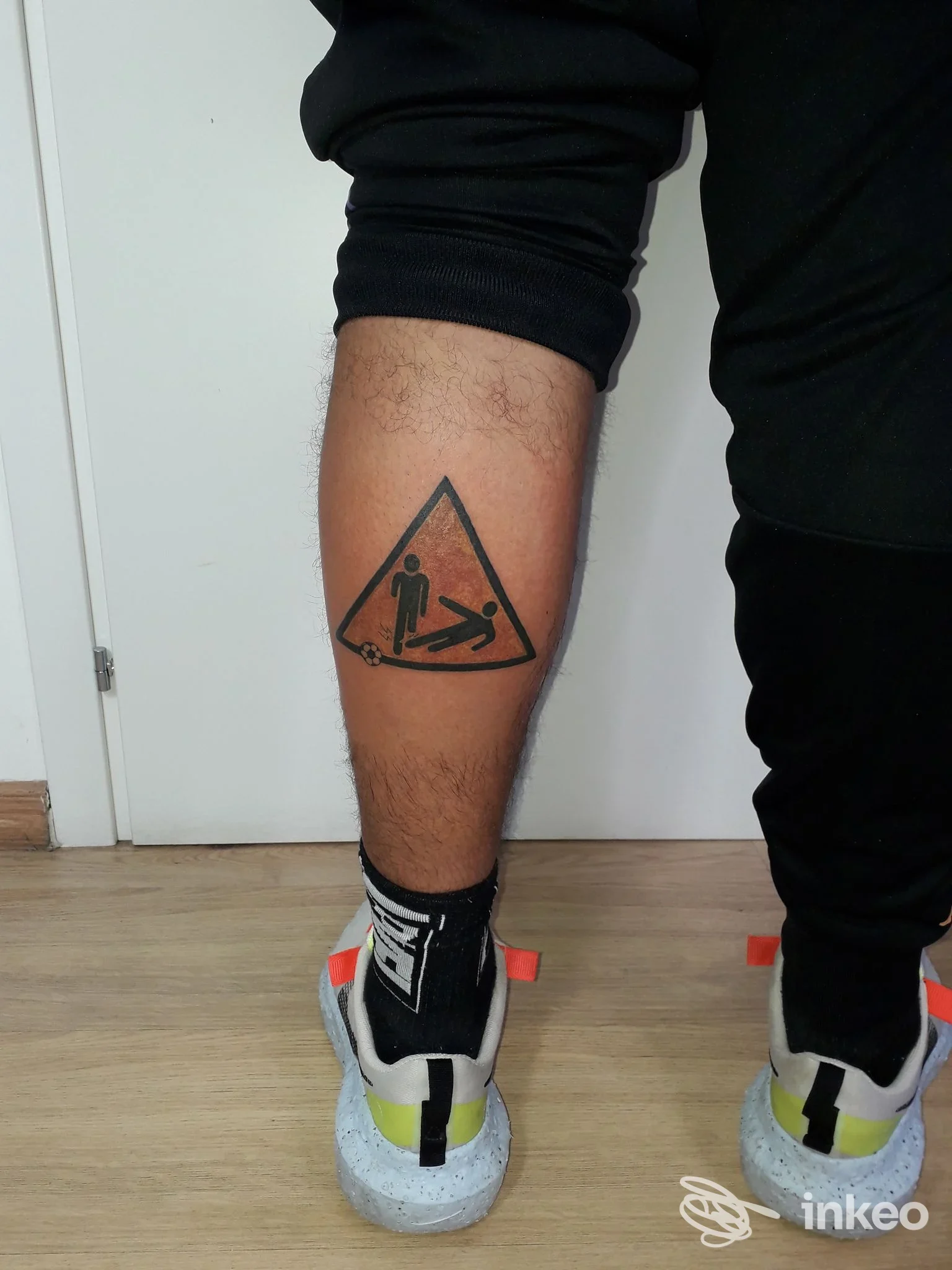 Tatuaggio sul polpaccio raffigurante un triangolo con una figura umana al suo interno e una seconda figura a terra, realizzato sulla pelle in bianco e nero, visibile senza altri elementi promozionali.