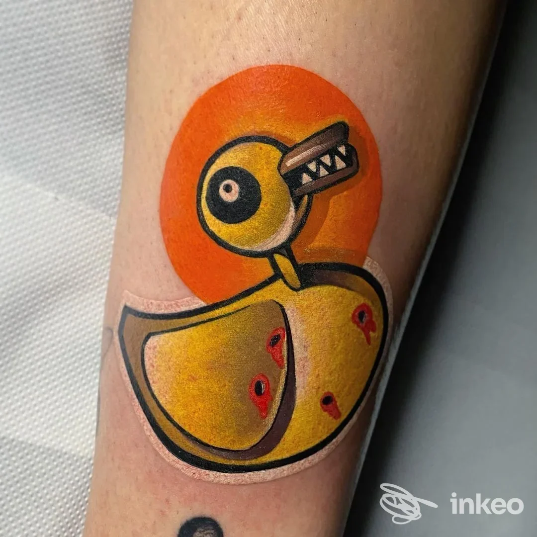 Tatuaggio cartoonesco su pelle: paperetta gialla con becco arancione, contorni neri su sfondo arancione, visibile sul braccio.