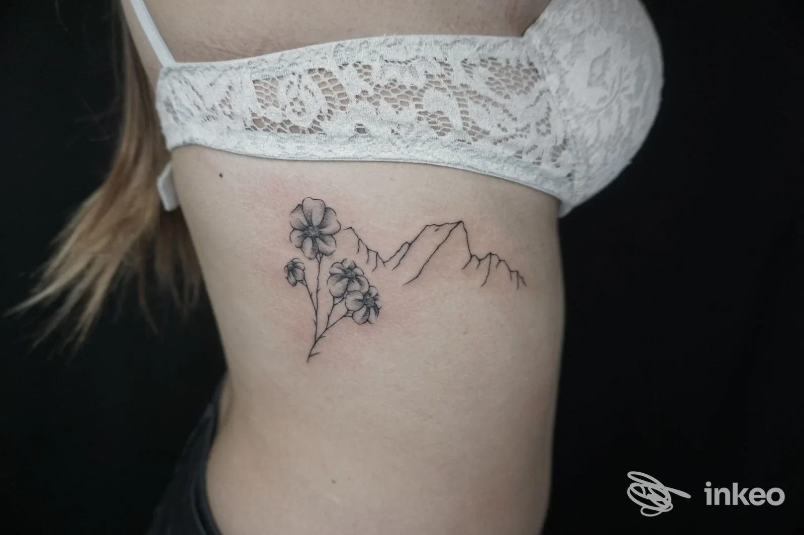 Tattoo sul fianco: fiori e catena di montagne in tratto sottile, disegno nero su pelle chiara, senza volto visibile.