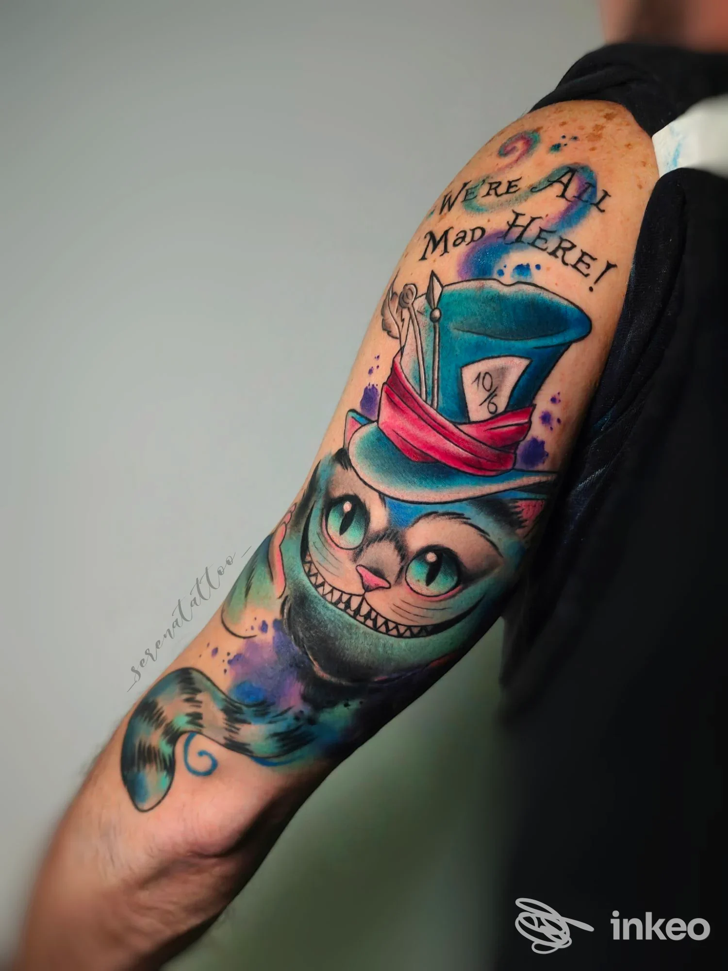 Tattoo colorato su avambraccio raffigurante una figura con cappello e una creatura, testo We are all mad here, visibile senza volto umano.