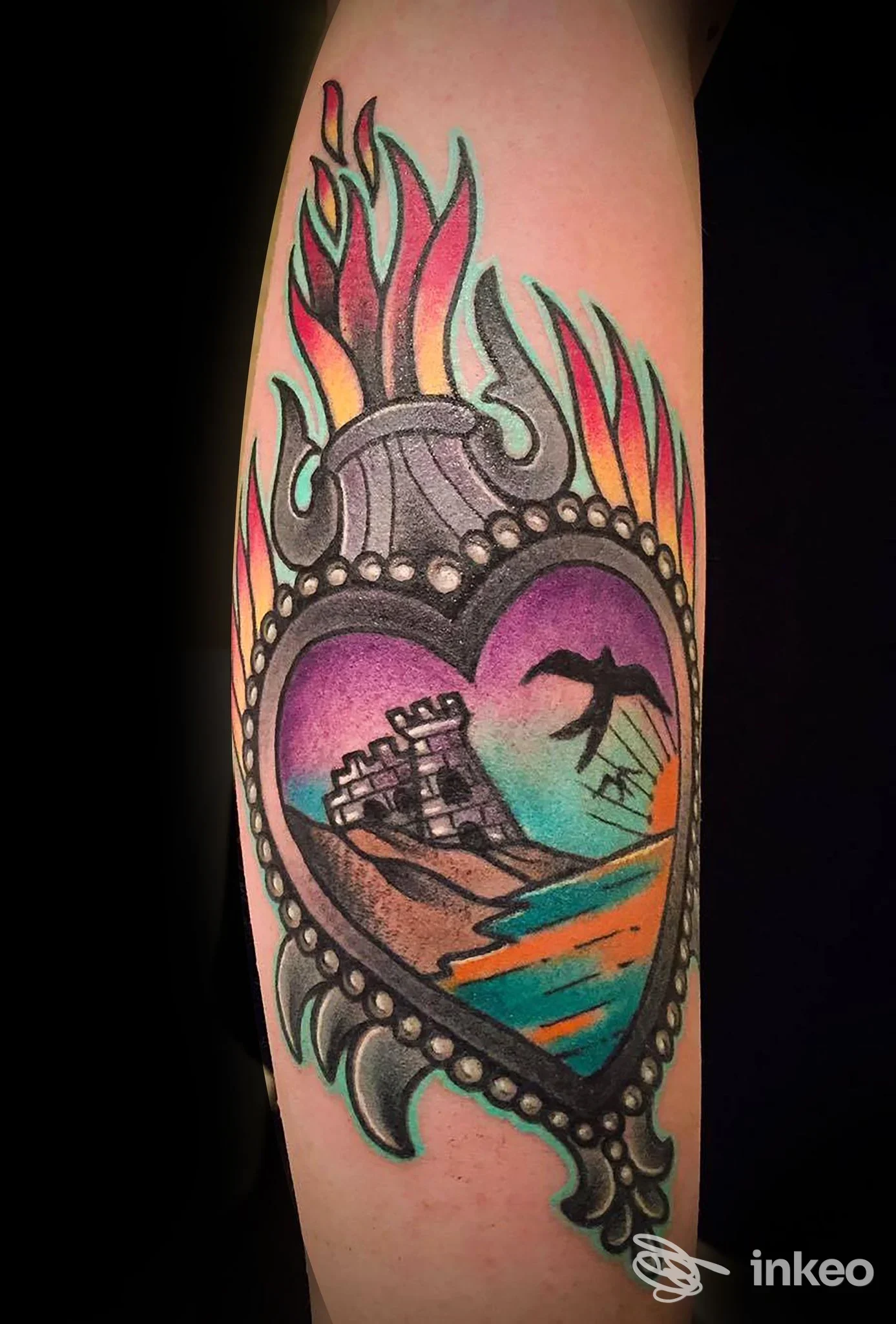 Tatuaggio a cuore colorato su pelle, con fiamme e scena di torre e mare all’interno, senza volto visibile.