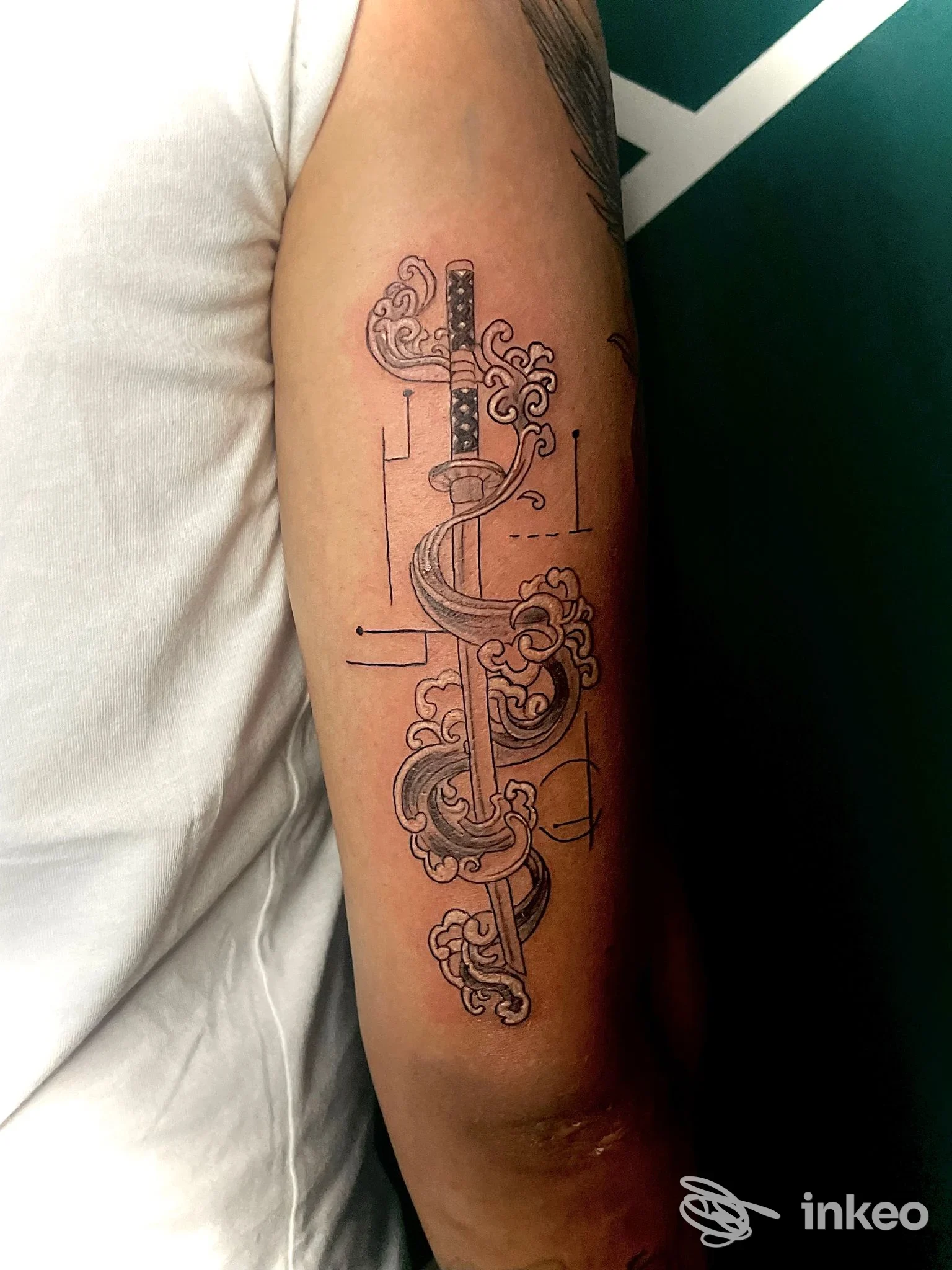 Tattoo su avambraccio sinistro raffigurante una spada ornata tra nuvole, con linee sottili e dettagli decorativi.