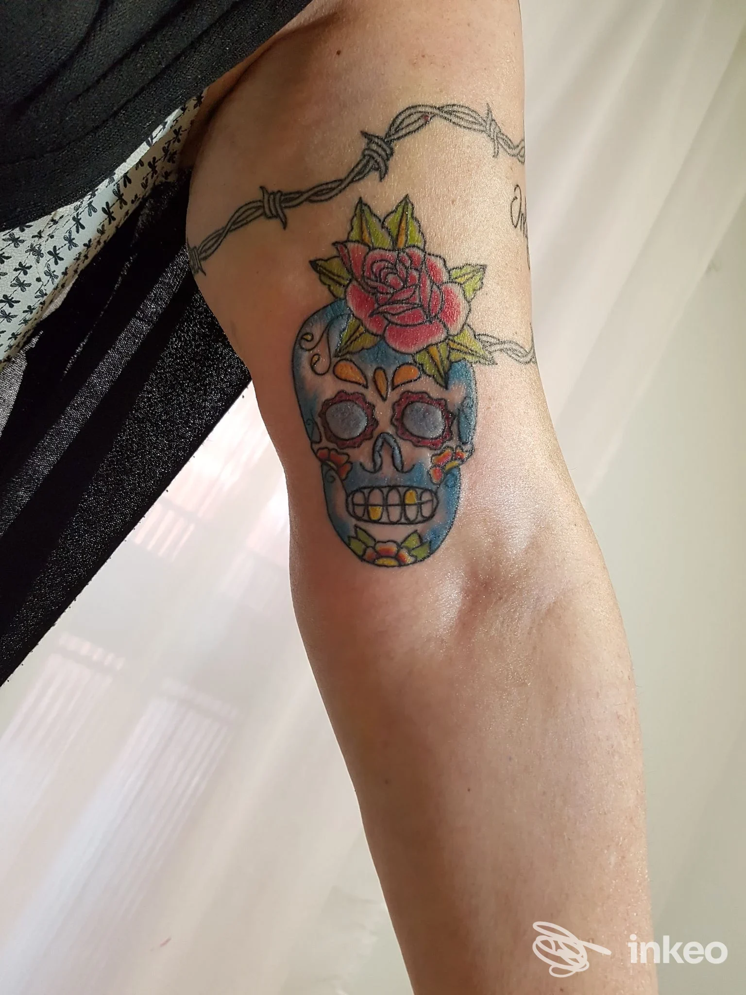 Tattoo a colori di una skull (calavera) con rosa sul braccio, con una cornicetta di filo spinato attorno al braccio; visibile pelle nuda e parte dell'arto superiore.