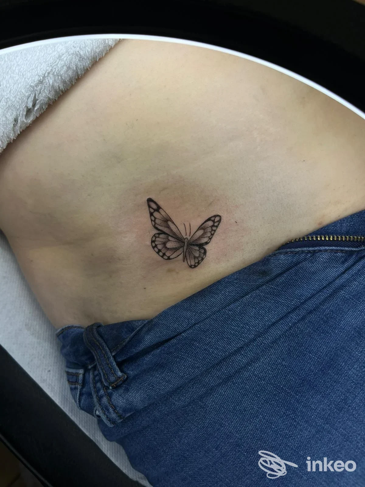 Piccolo tattoo di una farfalla in bianco e nero sul fianco sinistro vicino all'anca, visibile sulla pelle nuda.